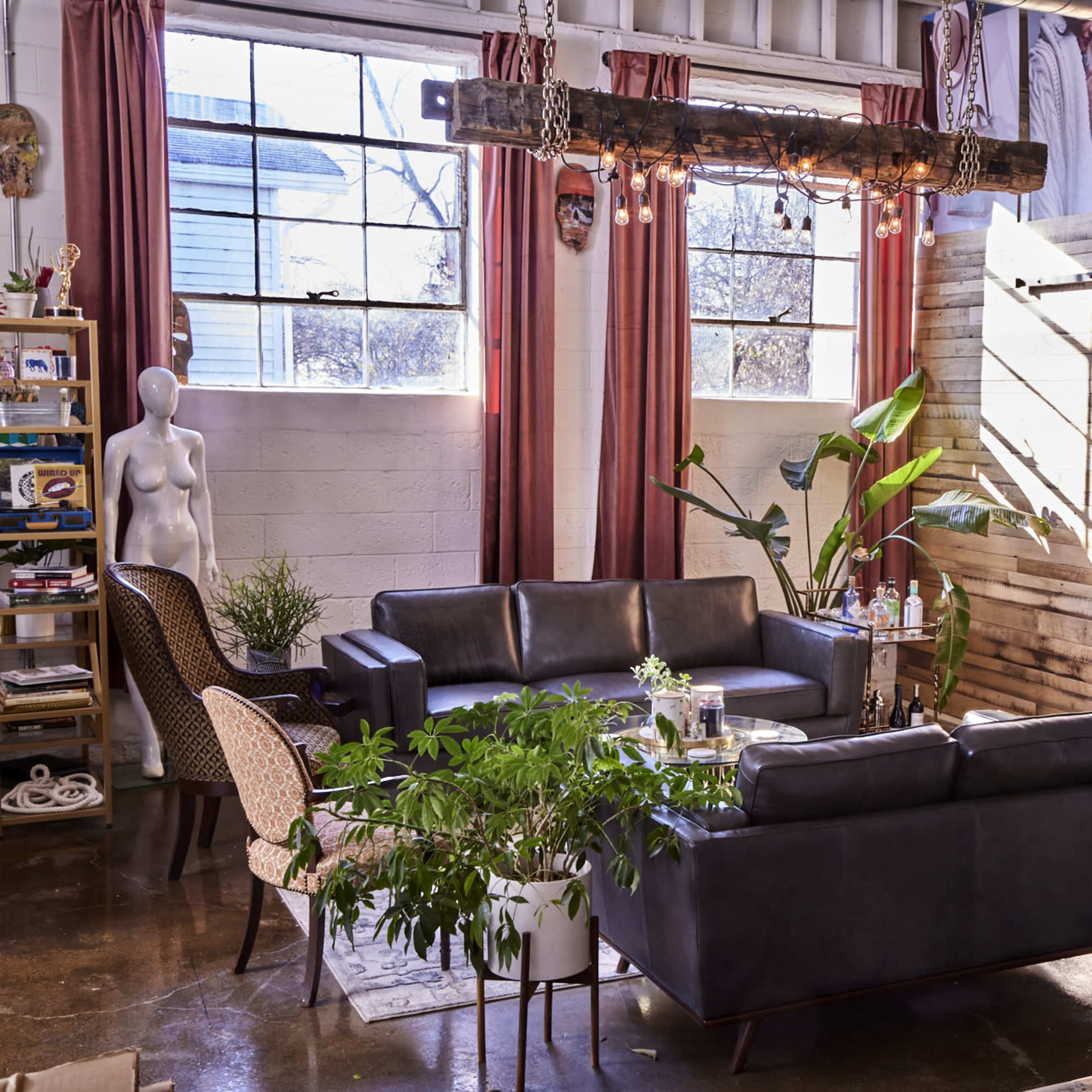 A modern living space with leather sofas, large windows, a wooden beam light fixture, potted plants, and a coffee station against a rustic wall.