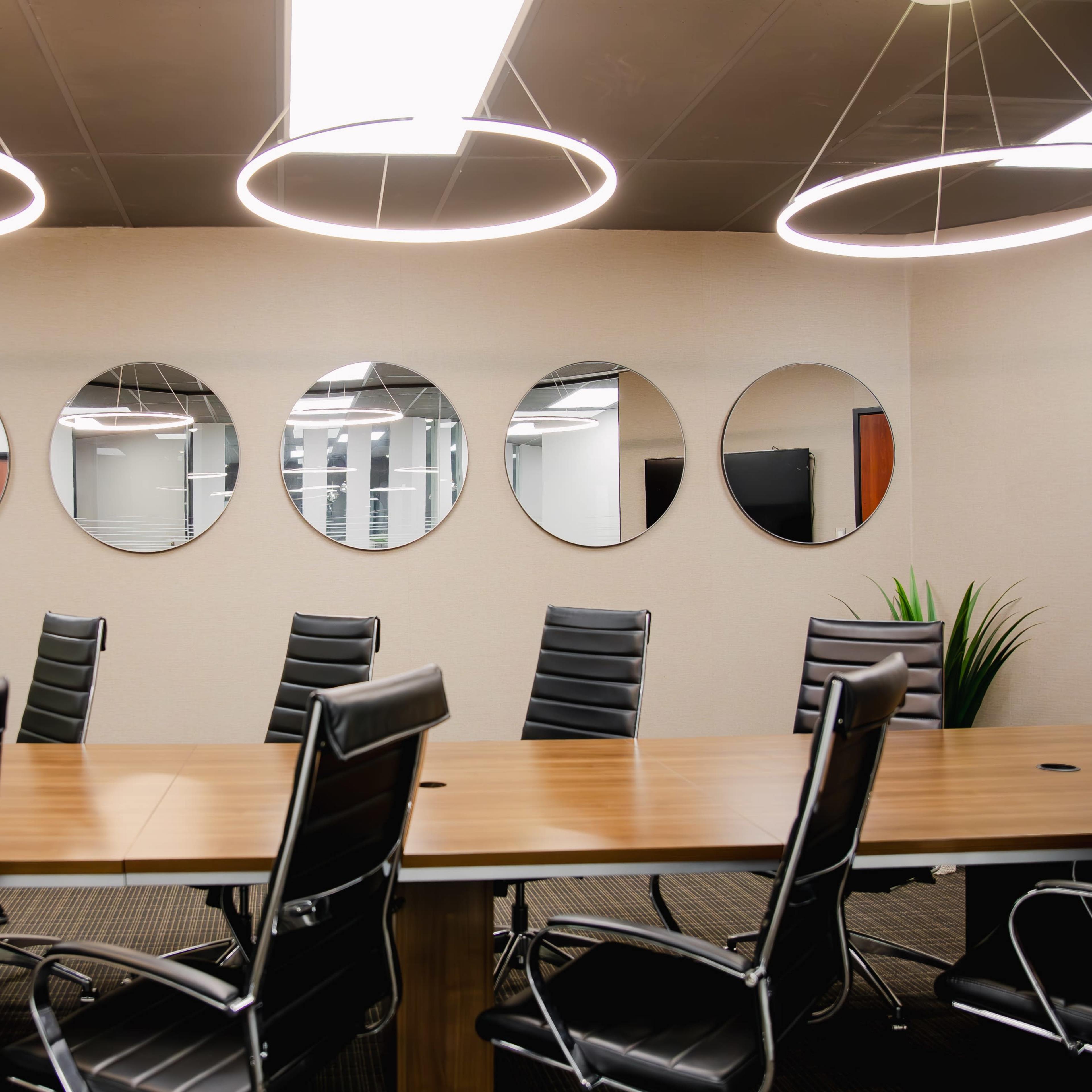 A modern conference room features a long wooden table surrounded by black leather chairs, with five round mirrors mounted on the wall and illuminated by circular pendant lights.