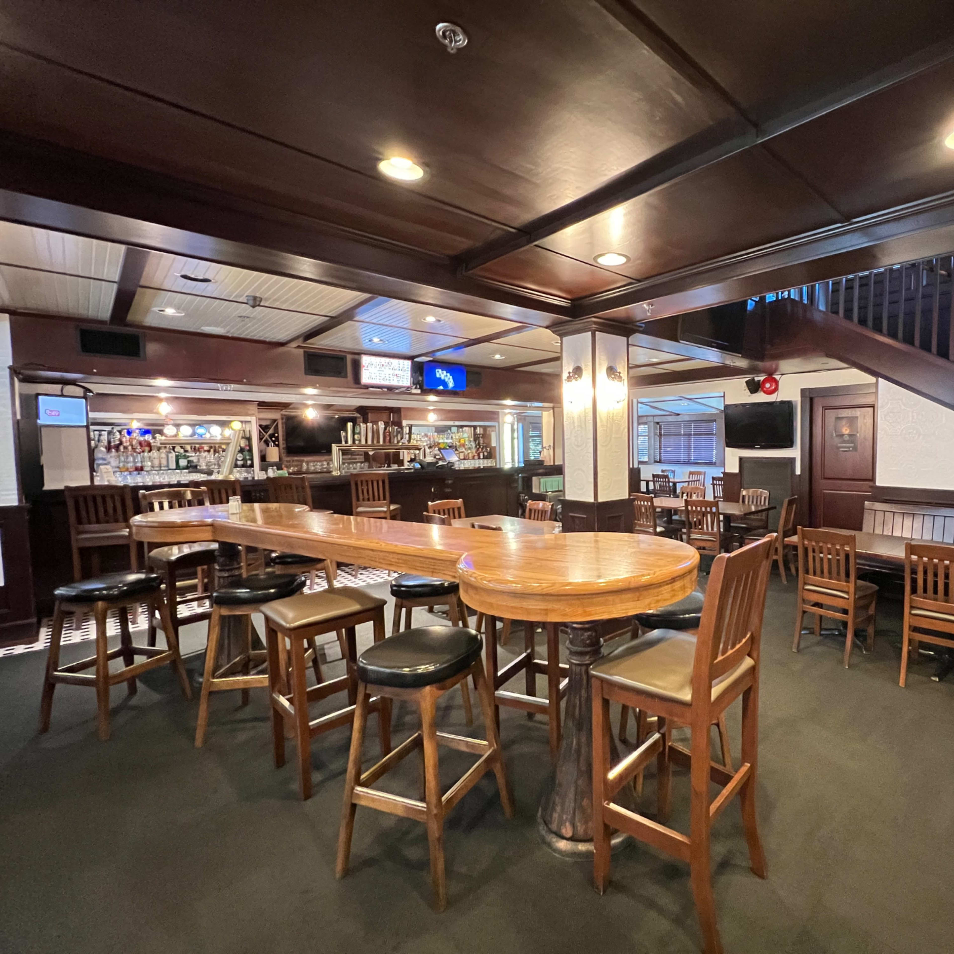 A spacious, dimly lit bar area with wooden furniture, a long table surrounded by high stools, and a staircase in the background.