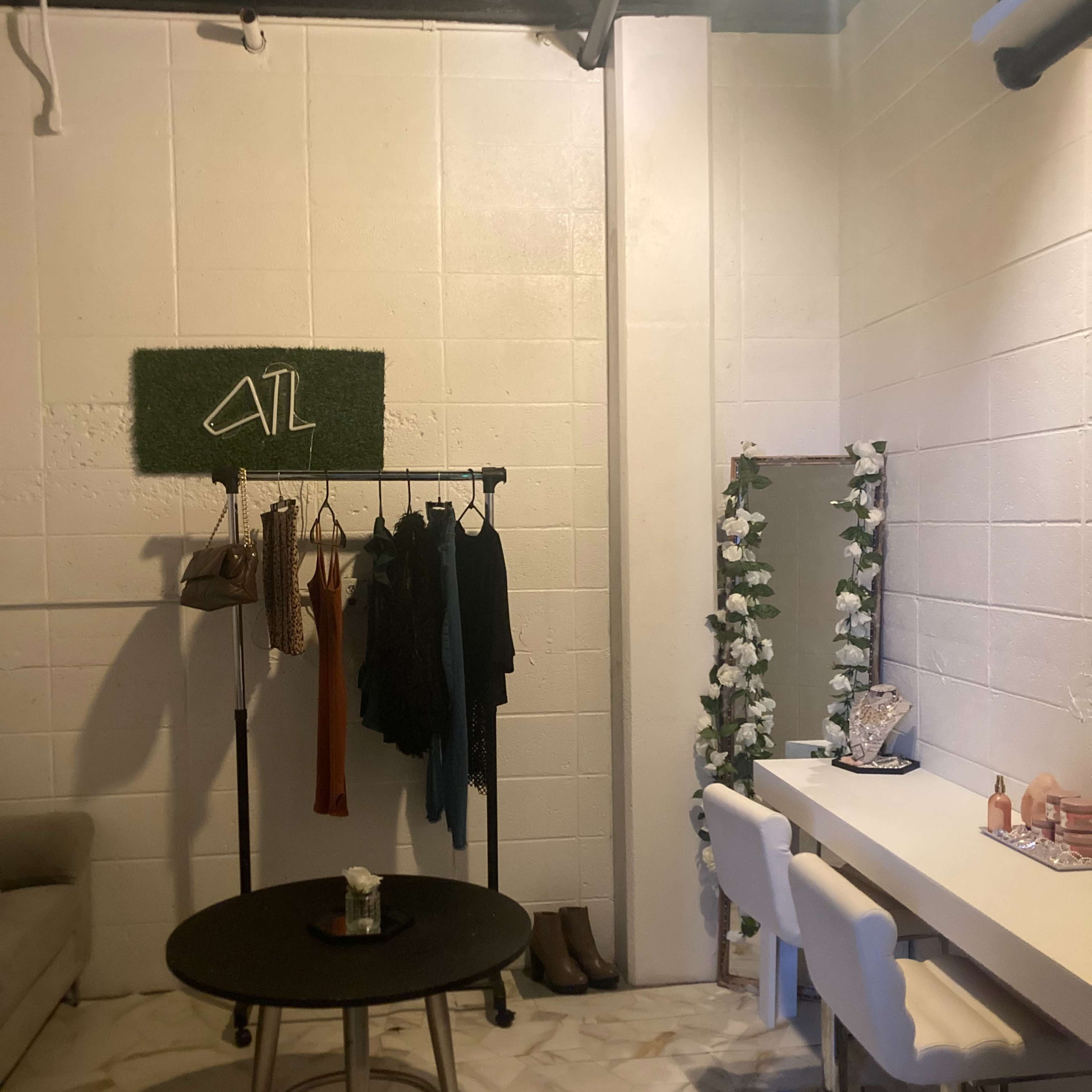A small room with a clothing rack displaying several garments, a round table in the center, and a vanity area with white stools and a mirror surrounded by flowers.