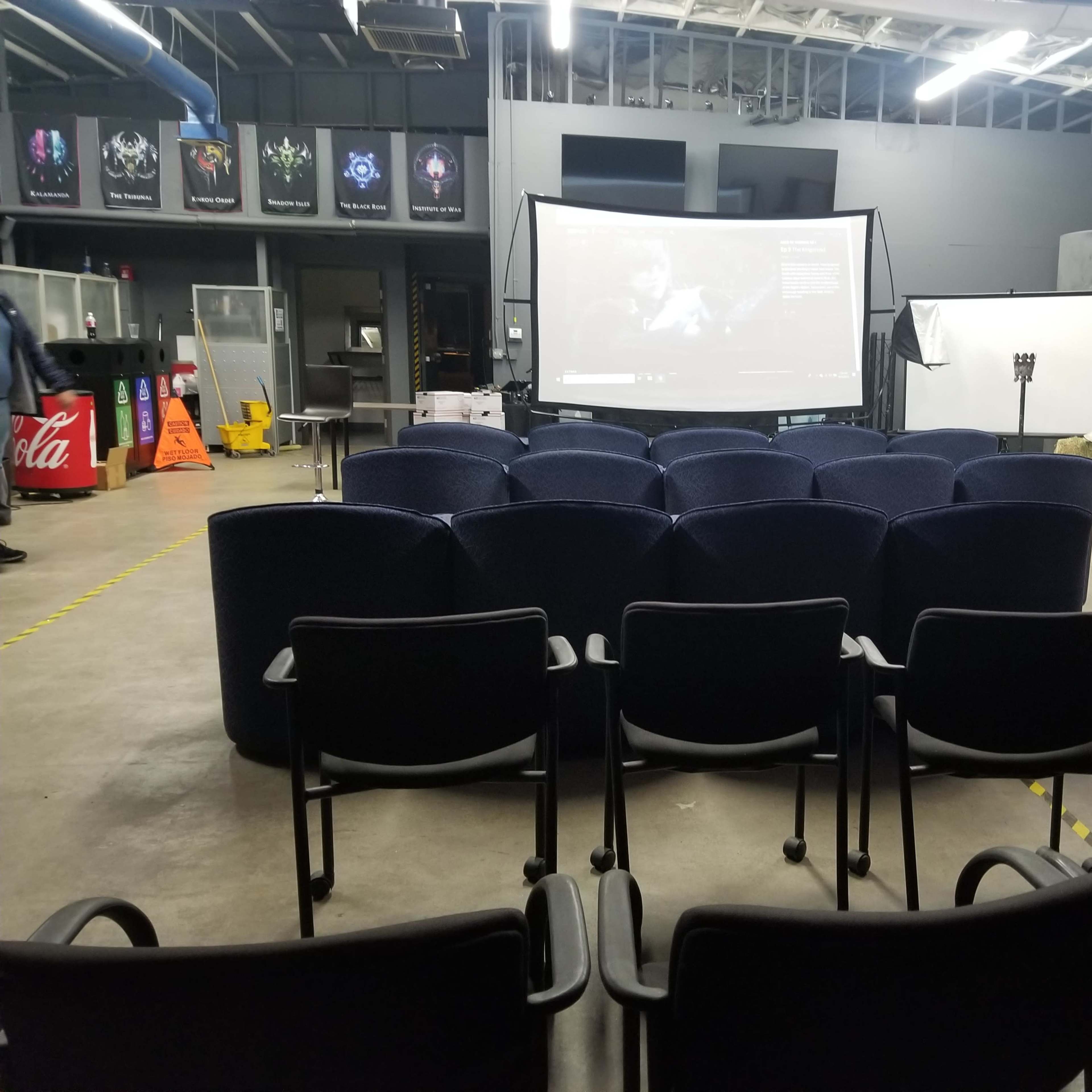 A room set up for a presentation, featuring rows of chairs facing a large screen displaying a video, with various equipment and posters on the walls.