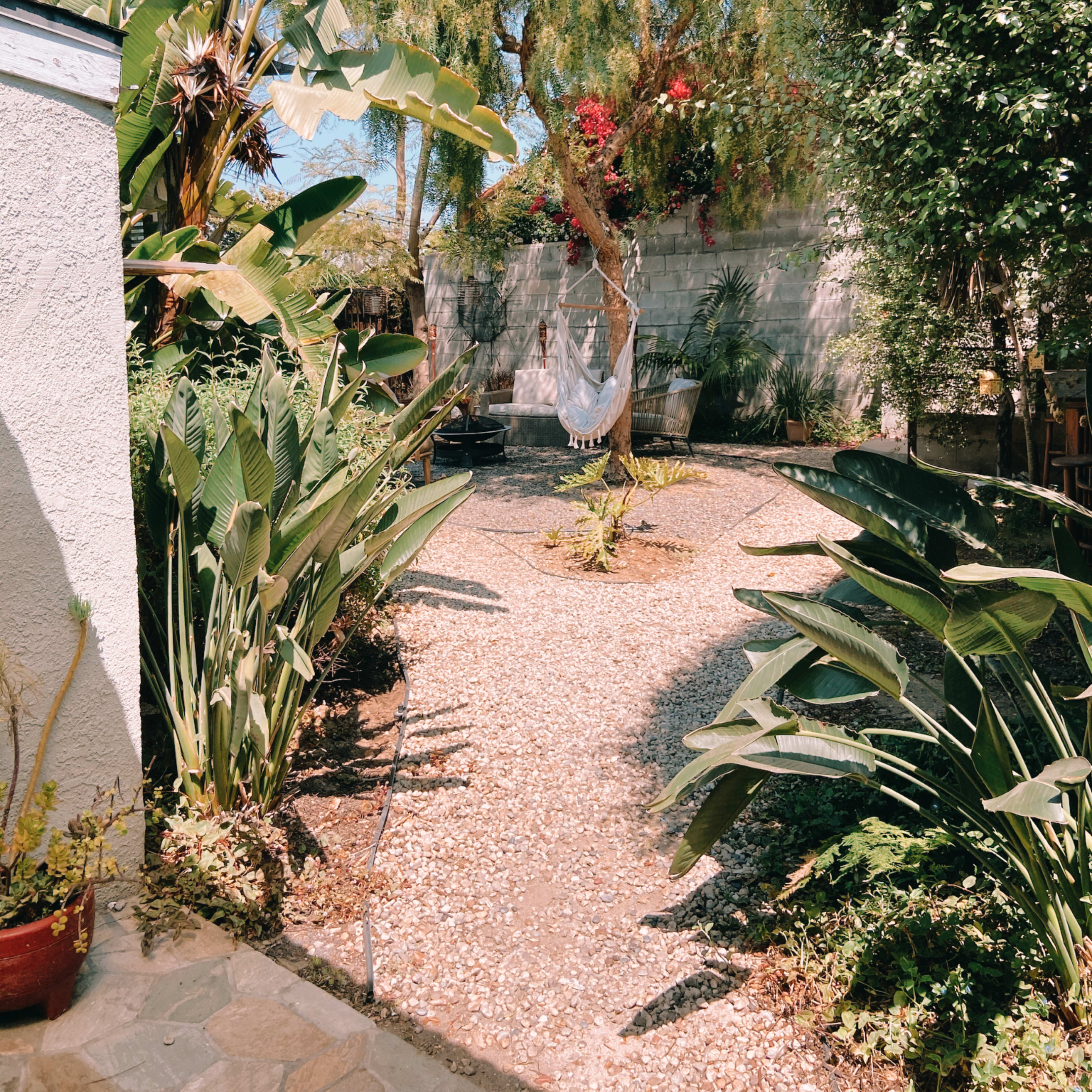 A gravel pathway leads through lush greenery and flowering plants in a sunny garden space, with a hammock hanging between two trees.