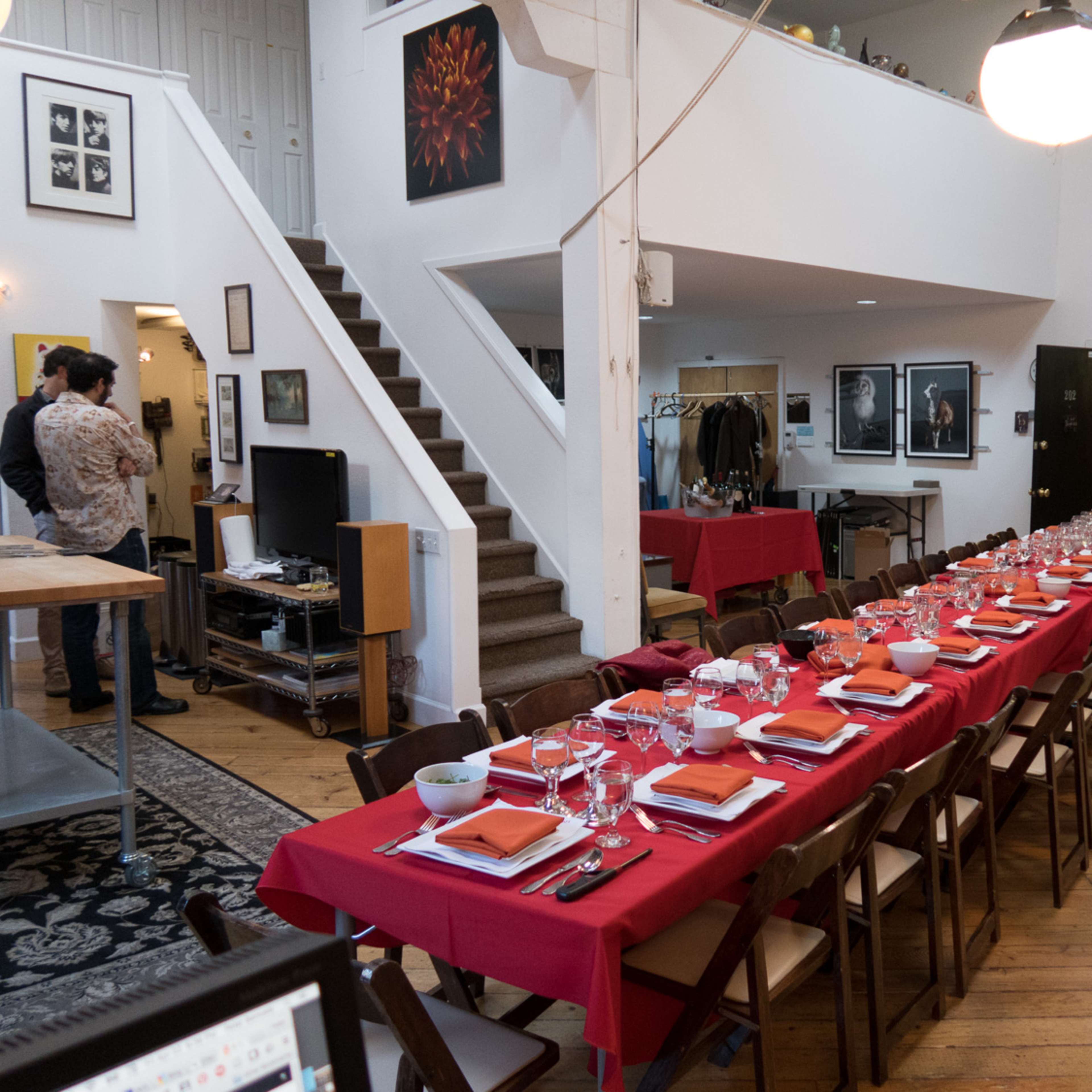 A long dining table with red tablecloths and orange napkins is set up in a spacious, well-lit room featuring stairs and various artworks on the walls.