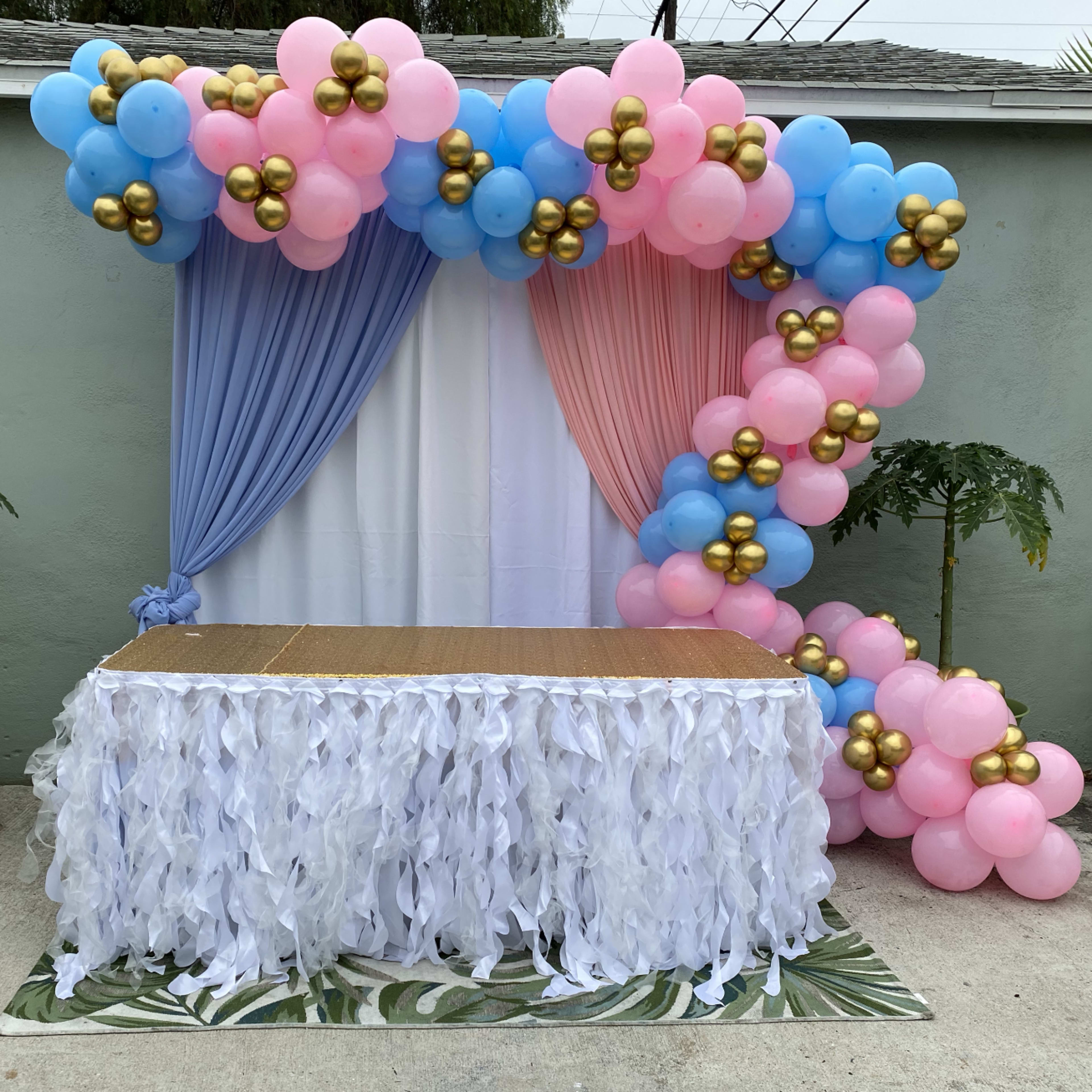 A decorated backdrop for an event, featuring blue, pink, and gold balloons arranged near a white table with a fringed tablecloth.