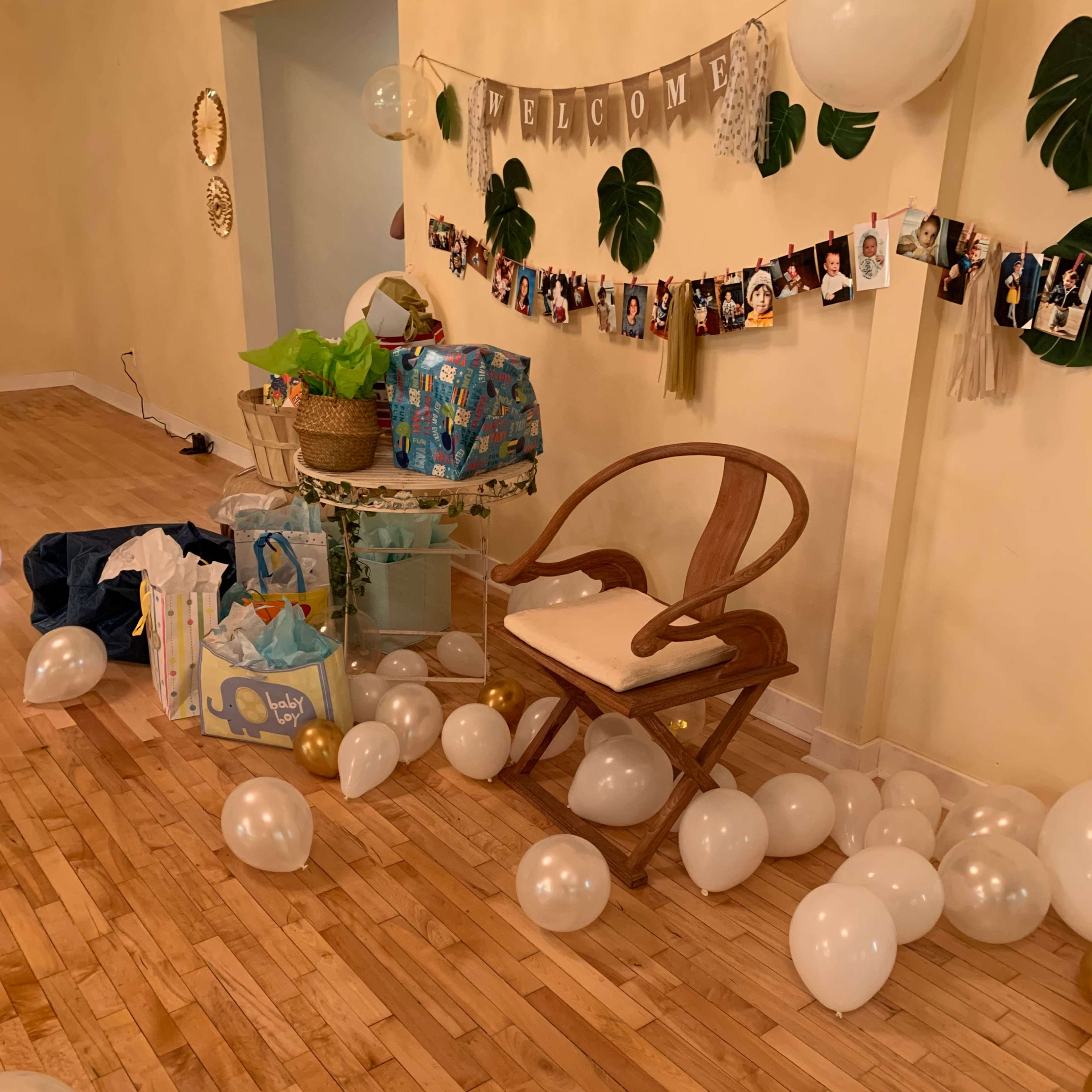 A room decorated for a celebration features a welcome banner, photos hanging on a string, a chair, and scattered white balloons on the wooden floor.