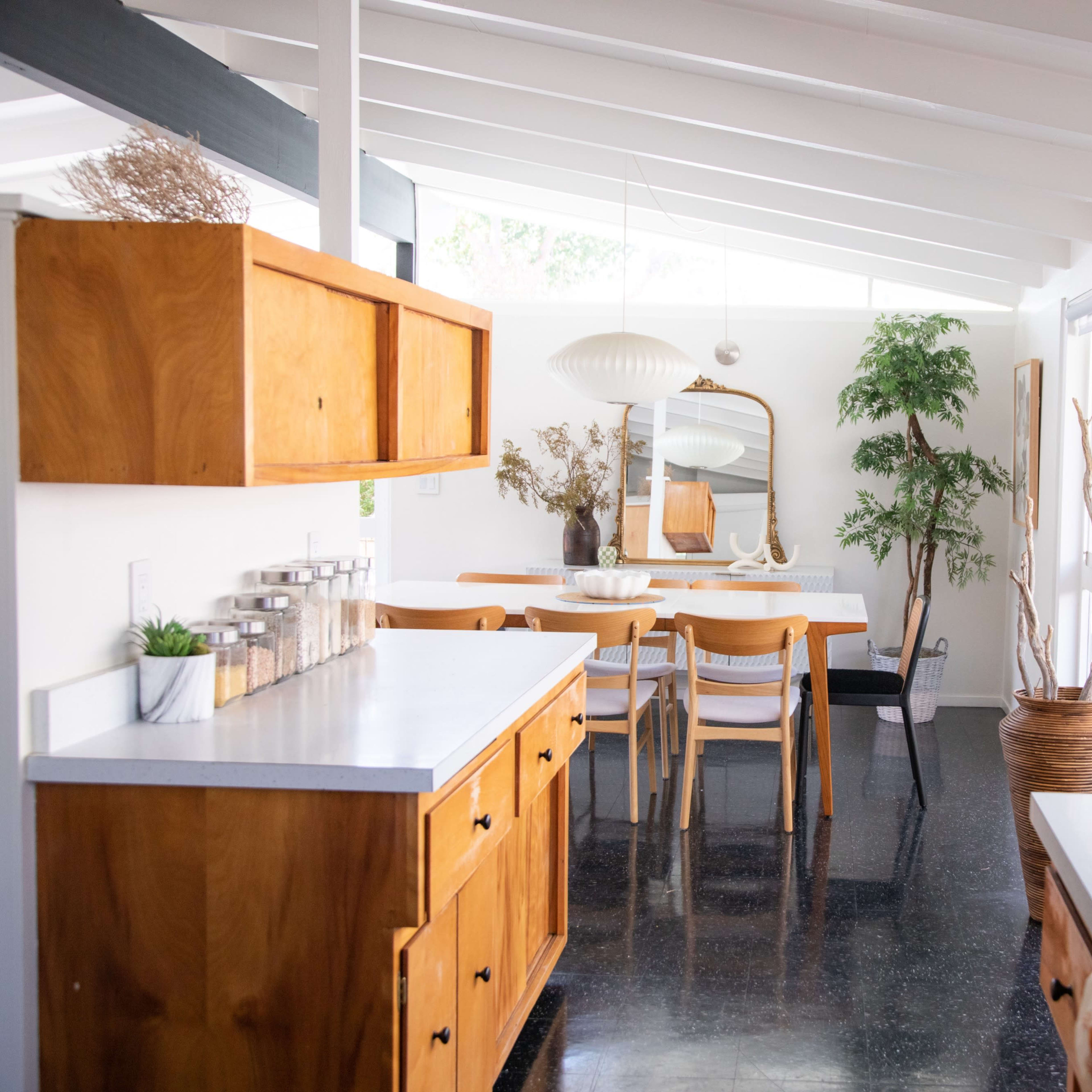 A bright and airy dining area features a white table surrounded by wooden chairs, with large windows letting in natural light and a view of greenery.