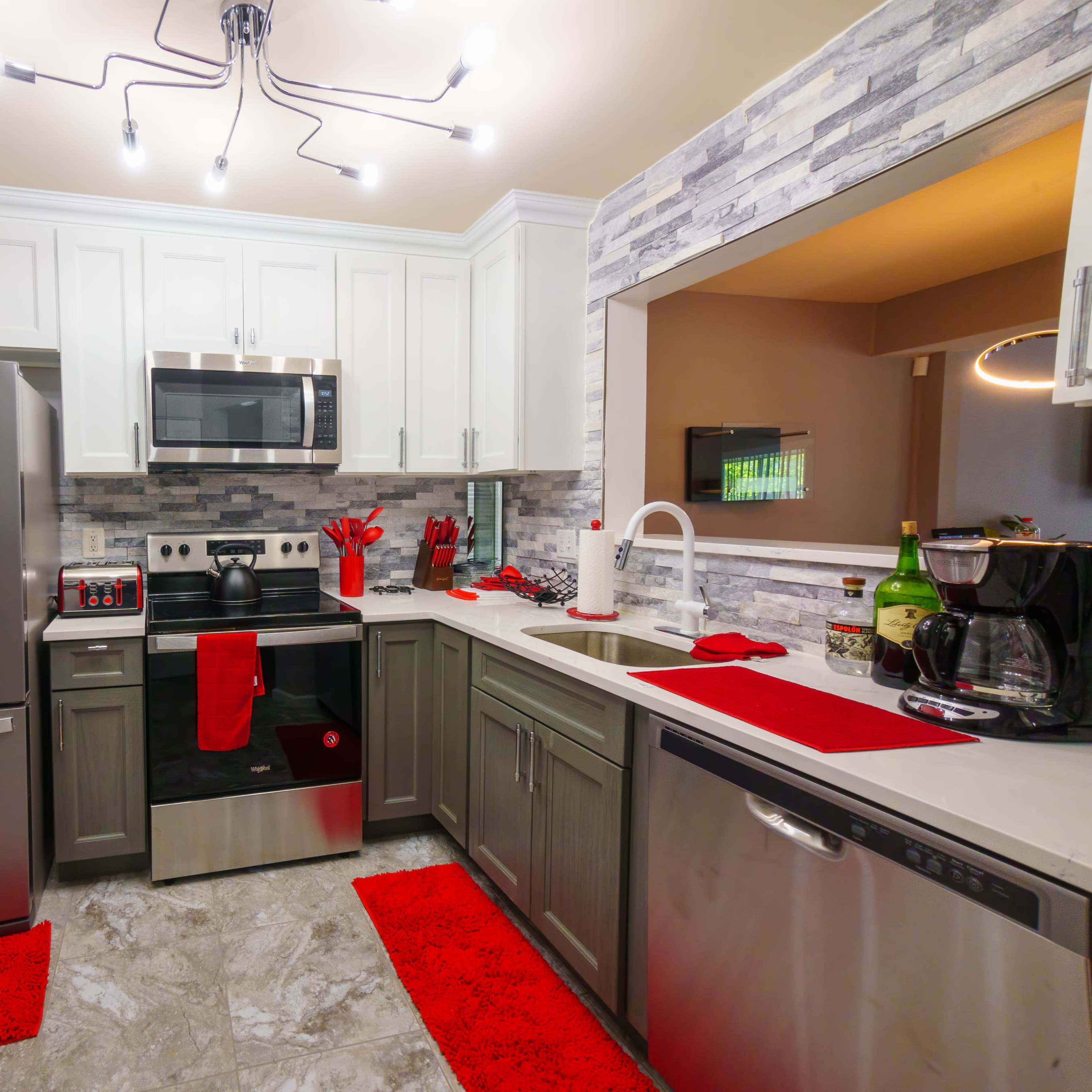 A modern kitchen featuring stainless steel appliances, white cabinets, and gray countertops, complemented by red accents on the dish towels and rugs.