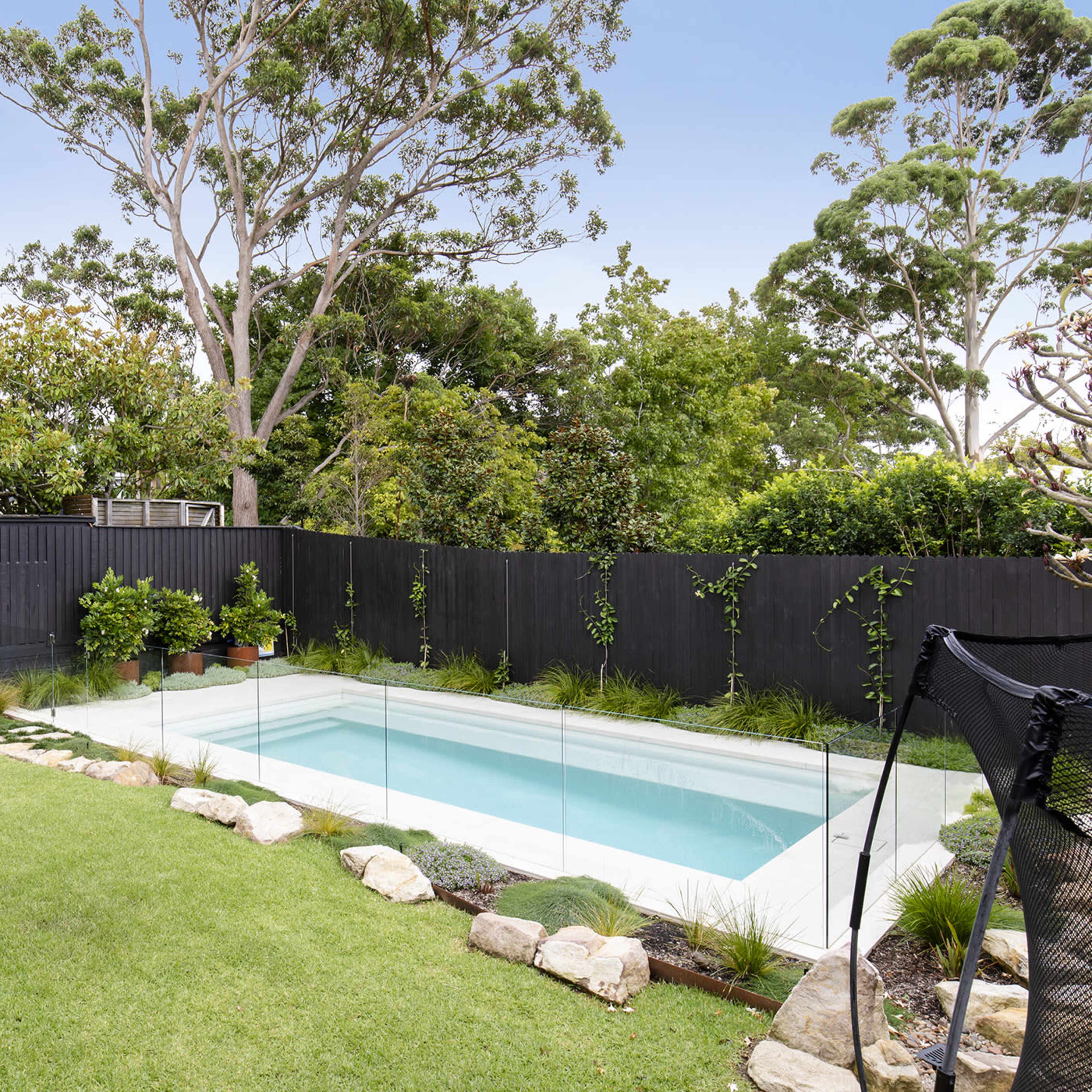 A clear swimming pool is surrounded by grass and decorative stones in a landscaped backyard with trees and a black fence.