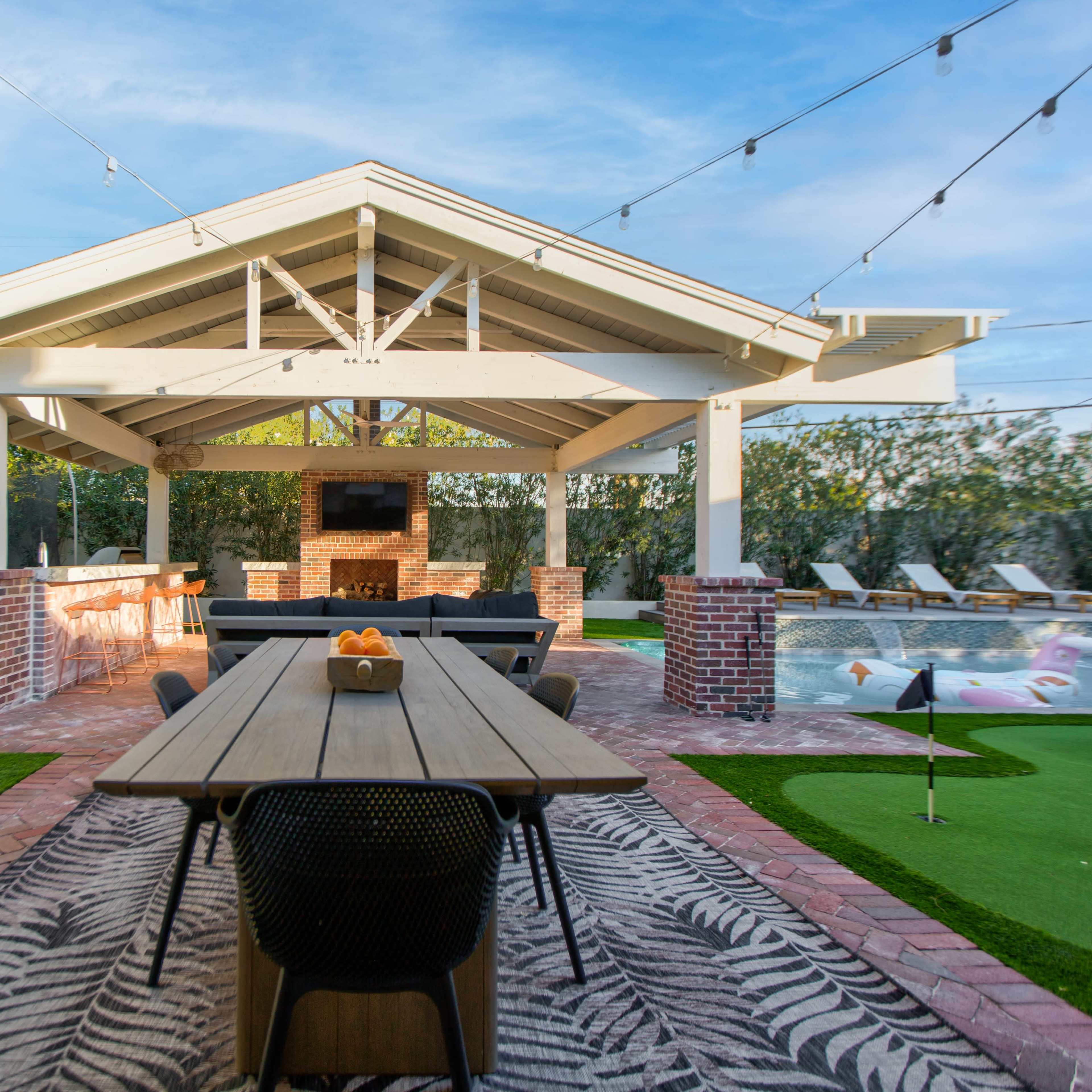 A backyard patio with a large table, a brick fireplace, and a swimming pool surrounded by green grass and landscaping under a clear sky.