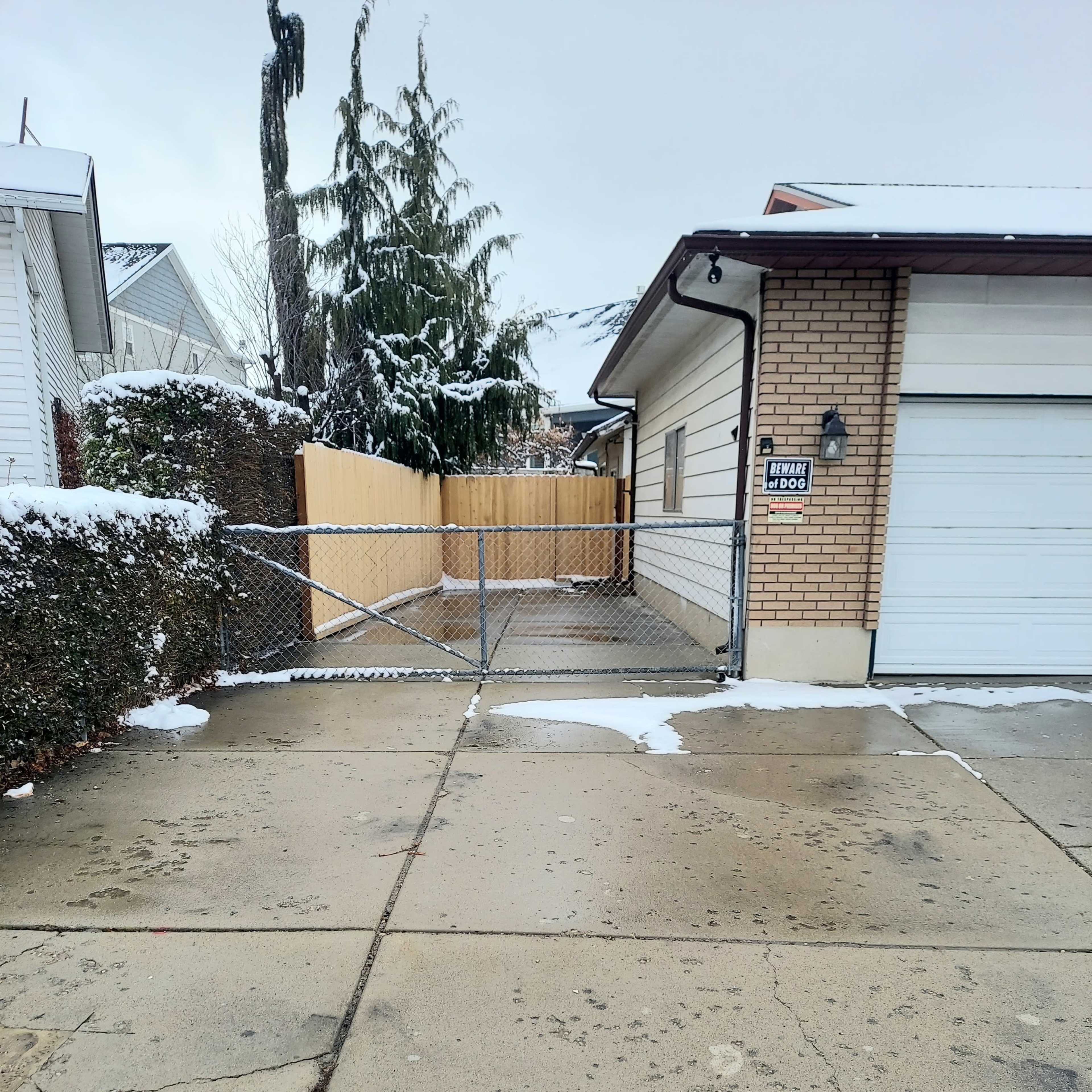 A snow-covered driveway with a chain-link fence leads to a house with a garage.