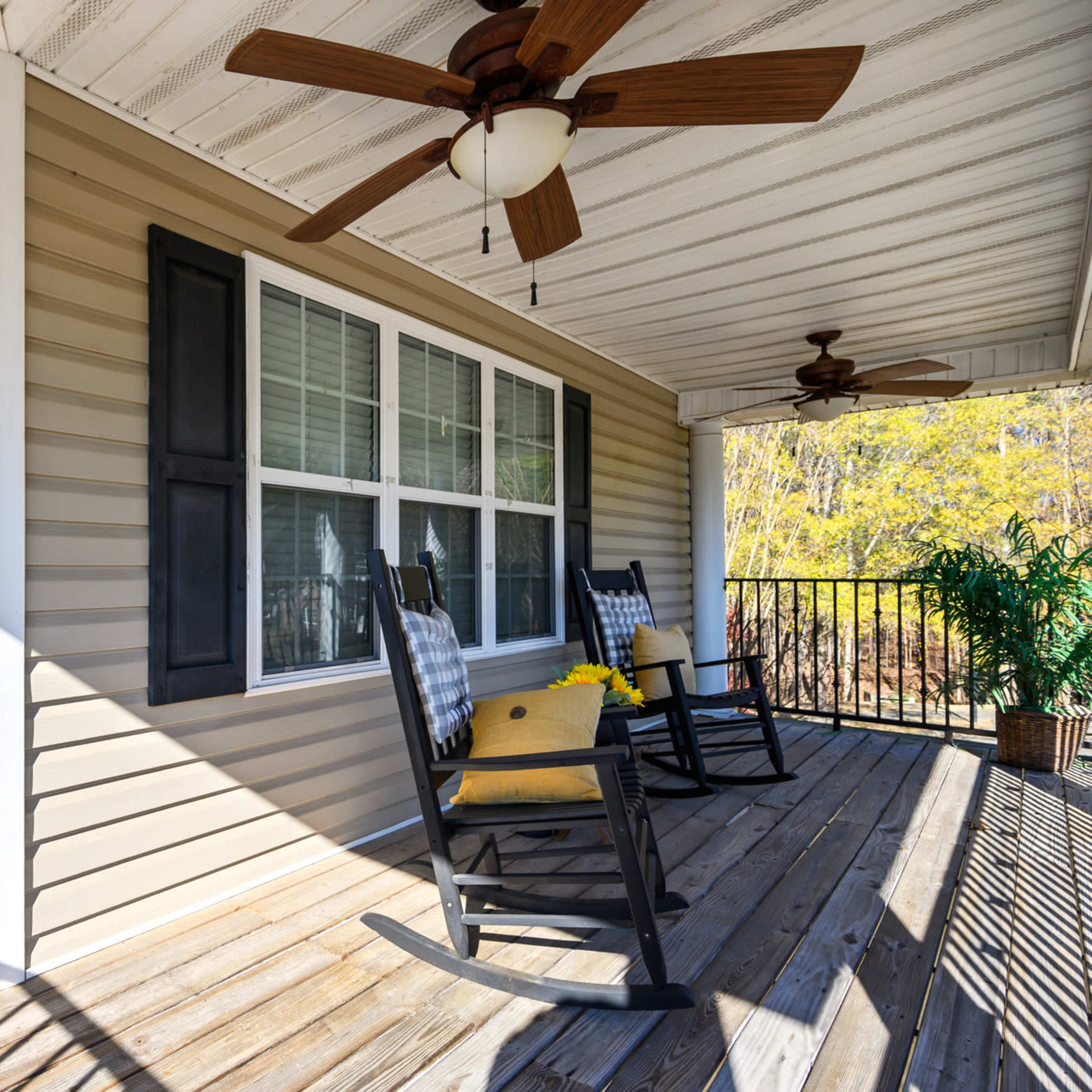 A covered porch featuring two rocking chairs, a houseplant, and large windows, with wooden decking and two ceiling fans overhead.