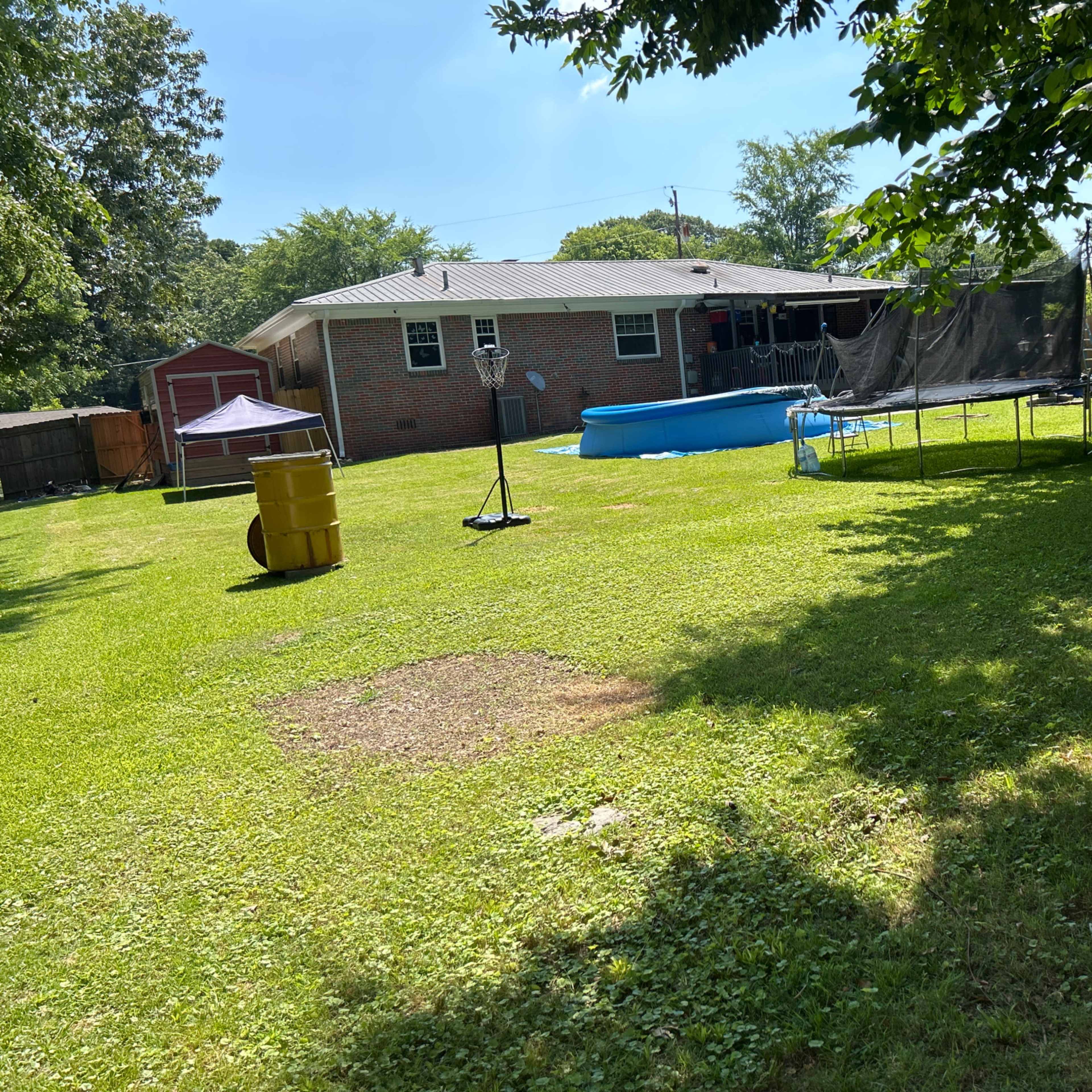A backyard featuring a swimming pool, a trampoline, a yellow barrel, and a brick house in the background.