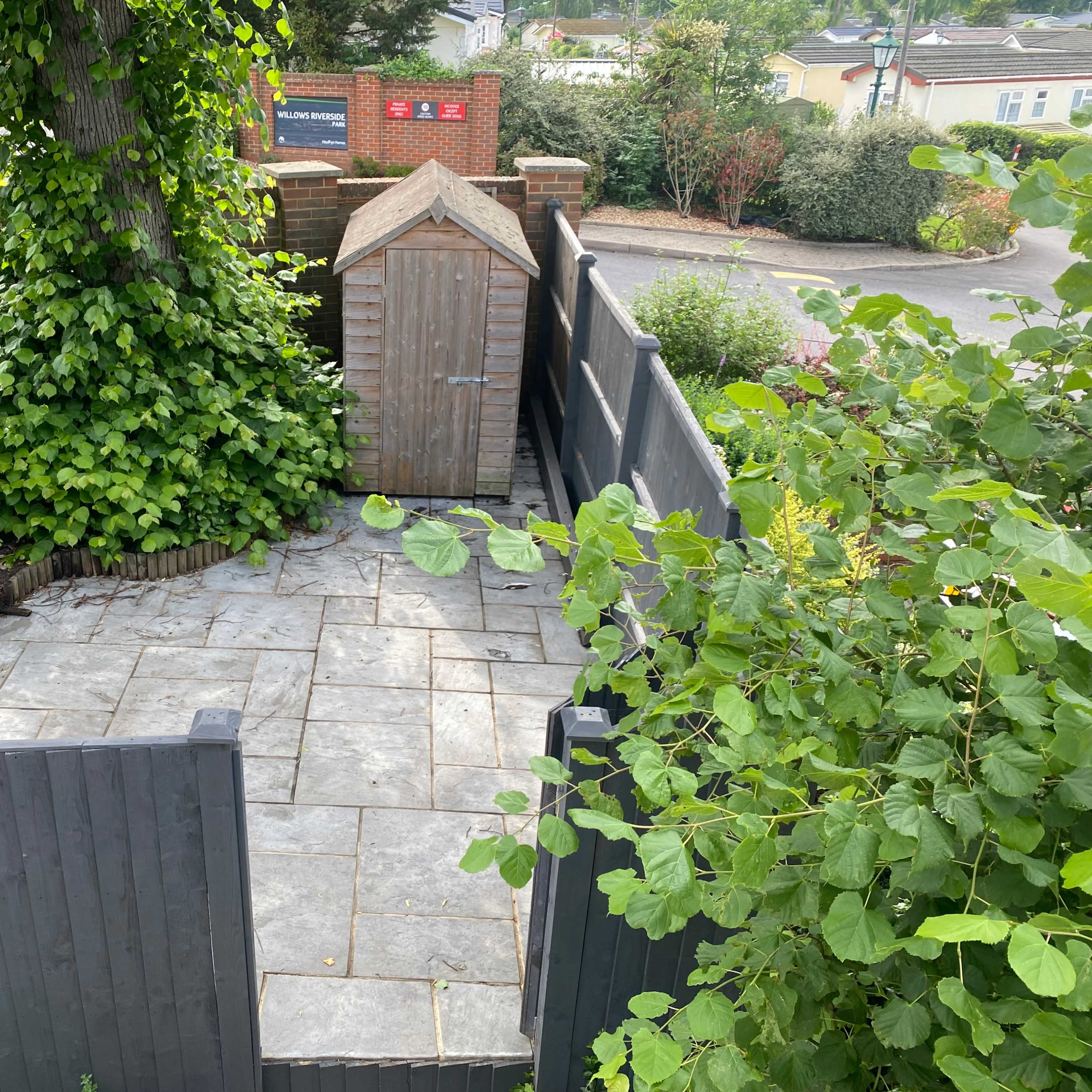 A fenced garden area with a stone patio, a small wooden shed, and surrounding greenery.