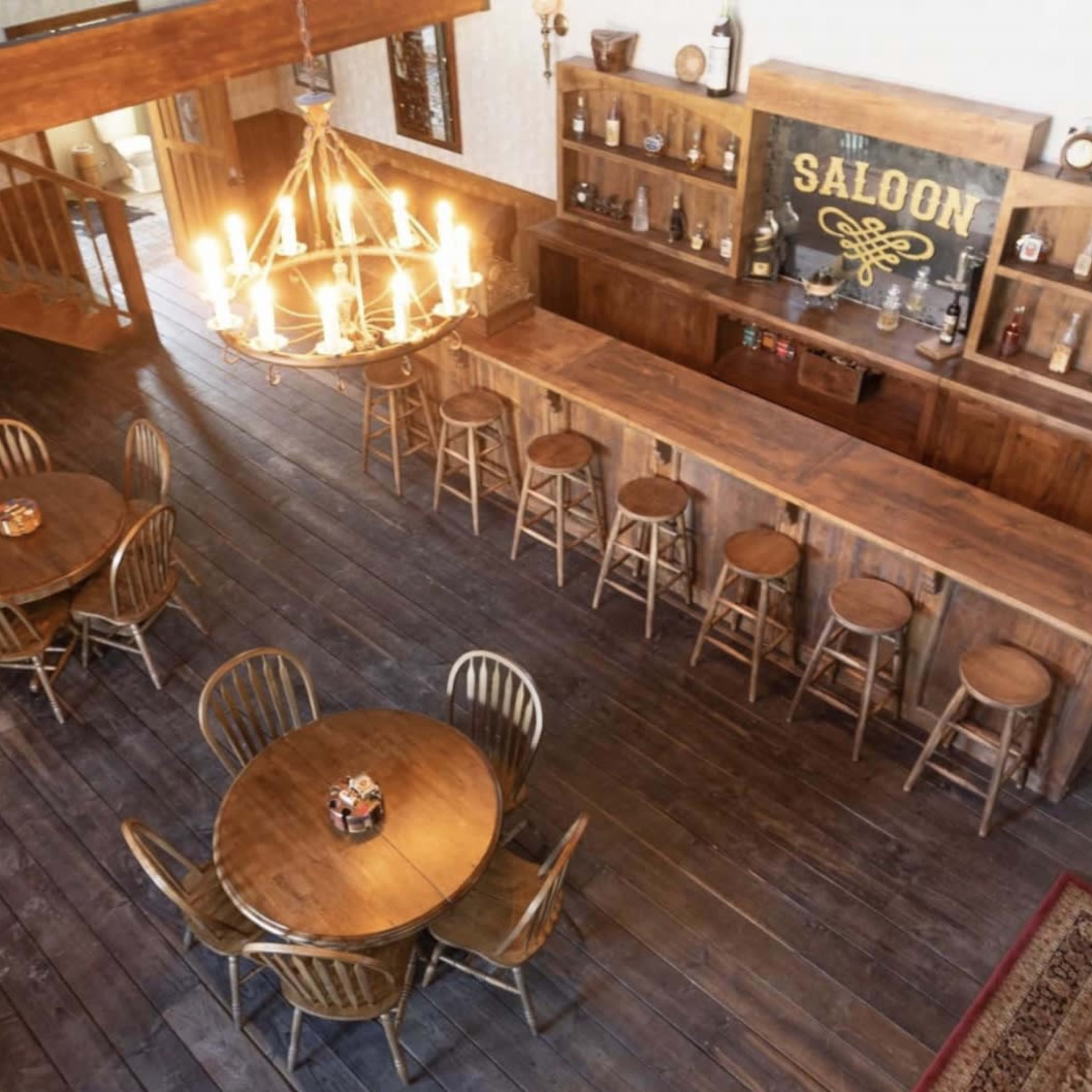 A rustic saloon interior featuring a wooden bar, stools, and round tables arranged beneath a chandelier.