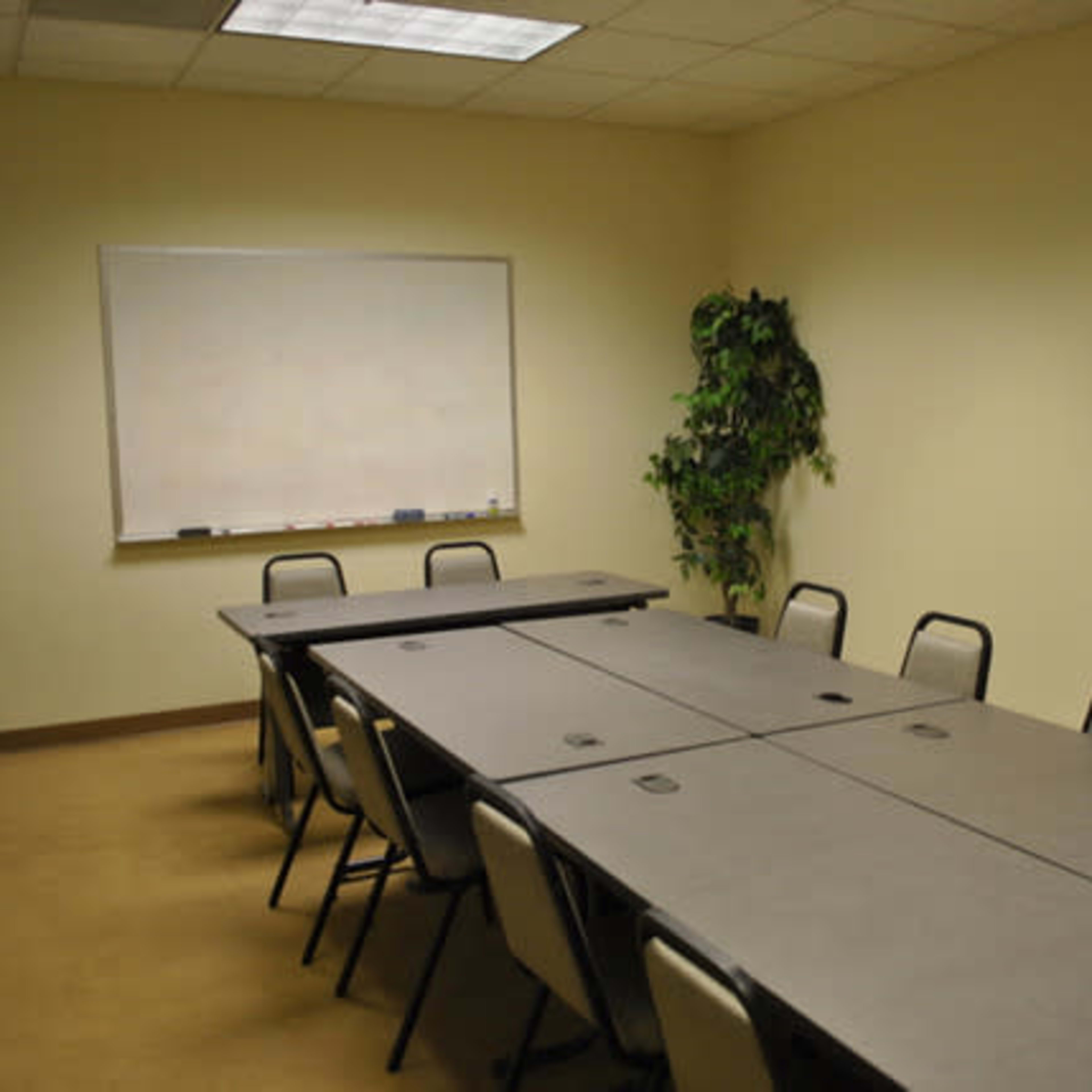 A sparse conference room features several rectangular tables arranged in a U-shape, with a whiteboard and a plant in the corner.