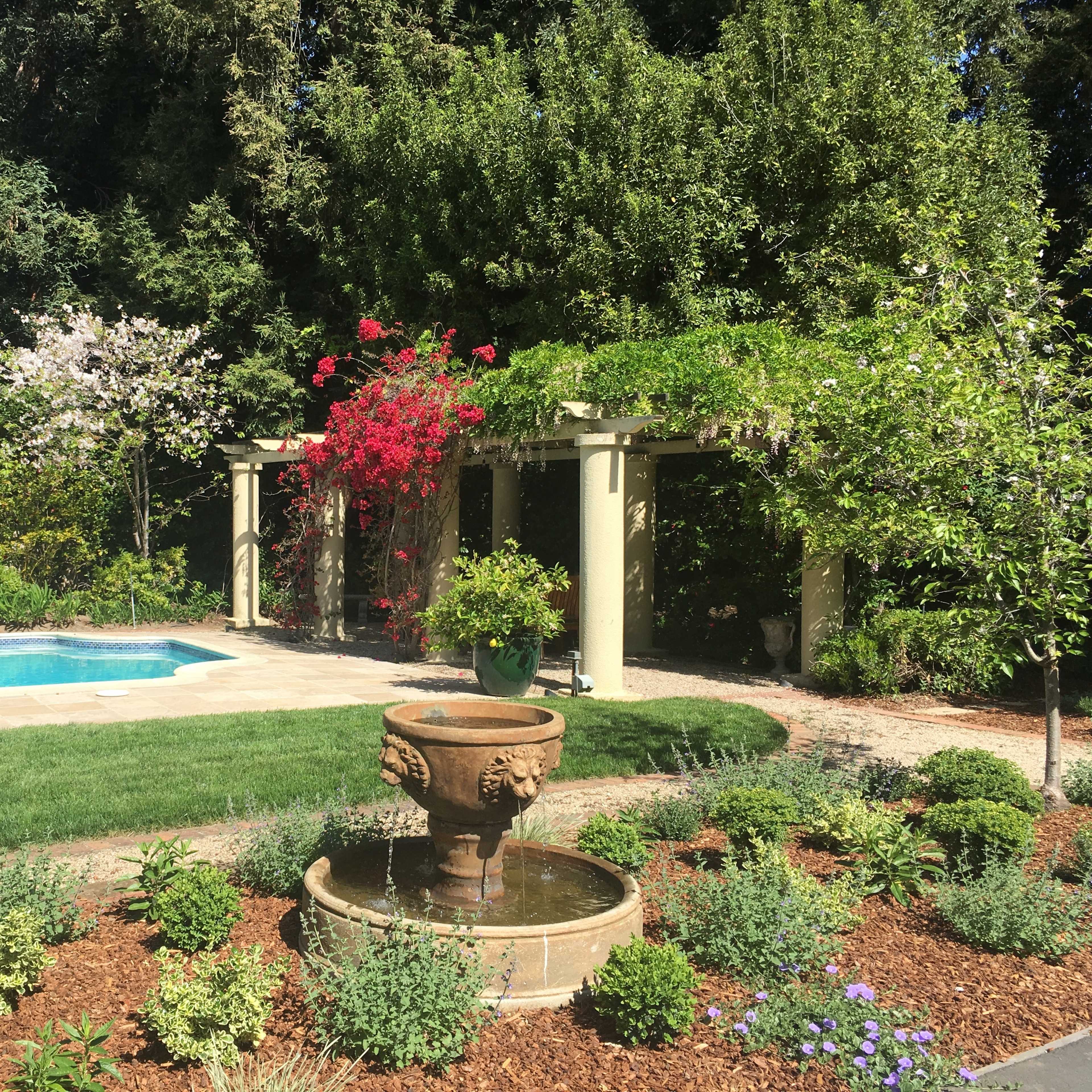 A landscaped garden with a pool, a stone fountain, and flowering plants, set against a backdrop of greenery.