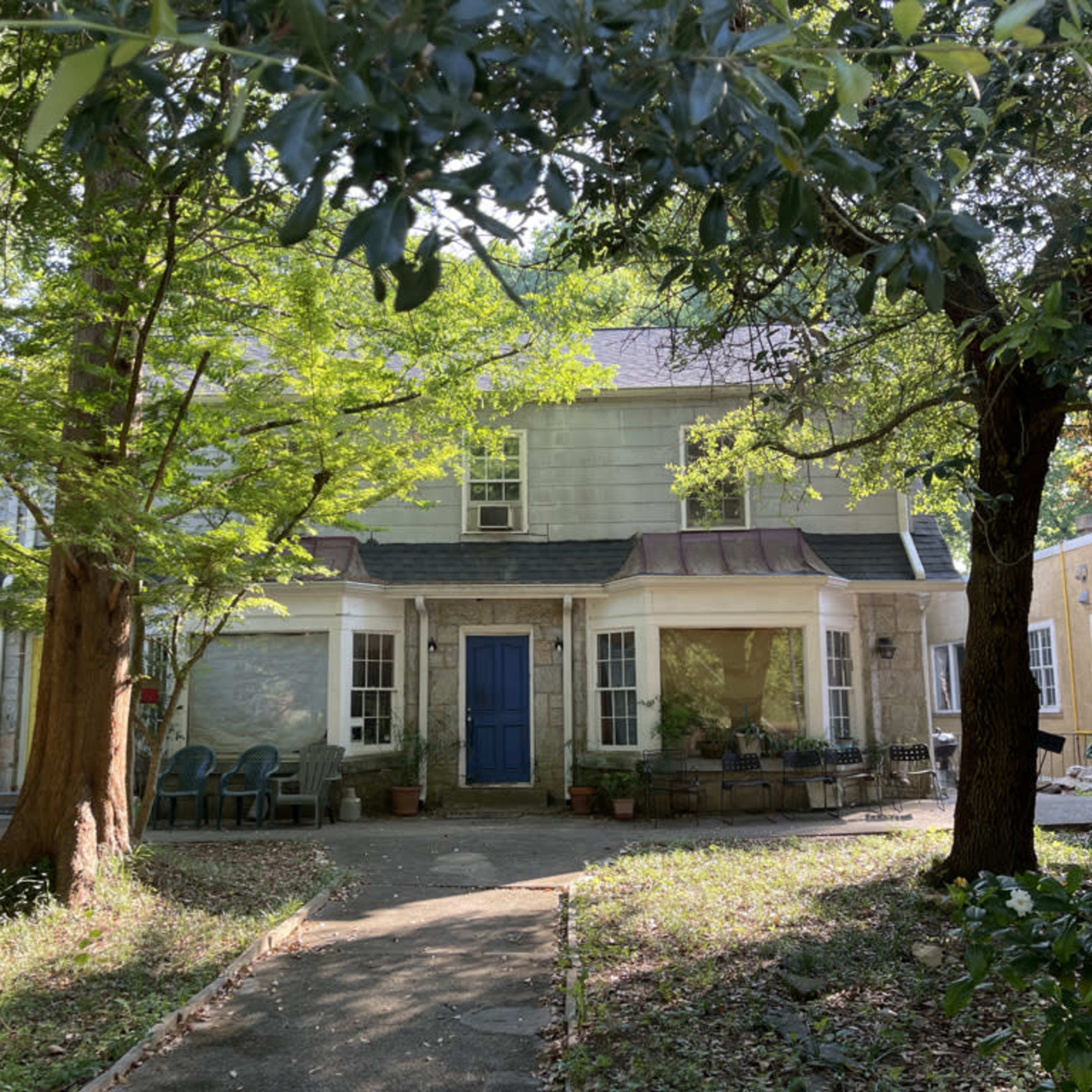A two-story house with a blue front door is partially obscured by trees and surrounded by greenery.