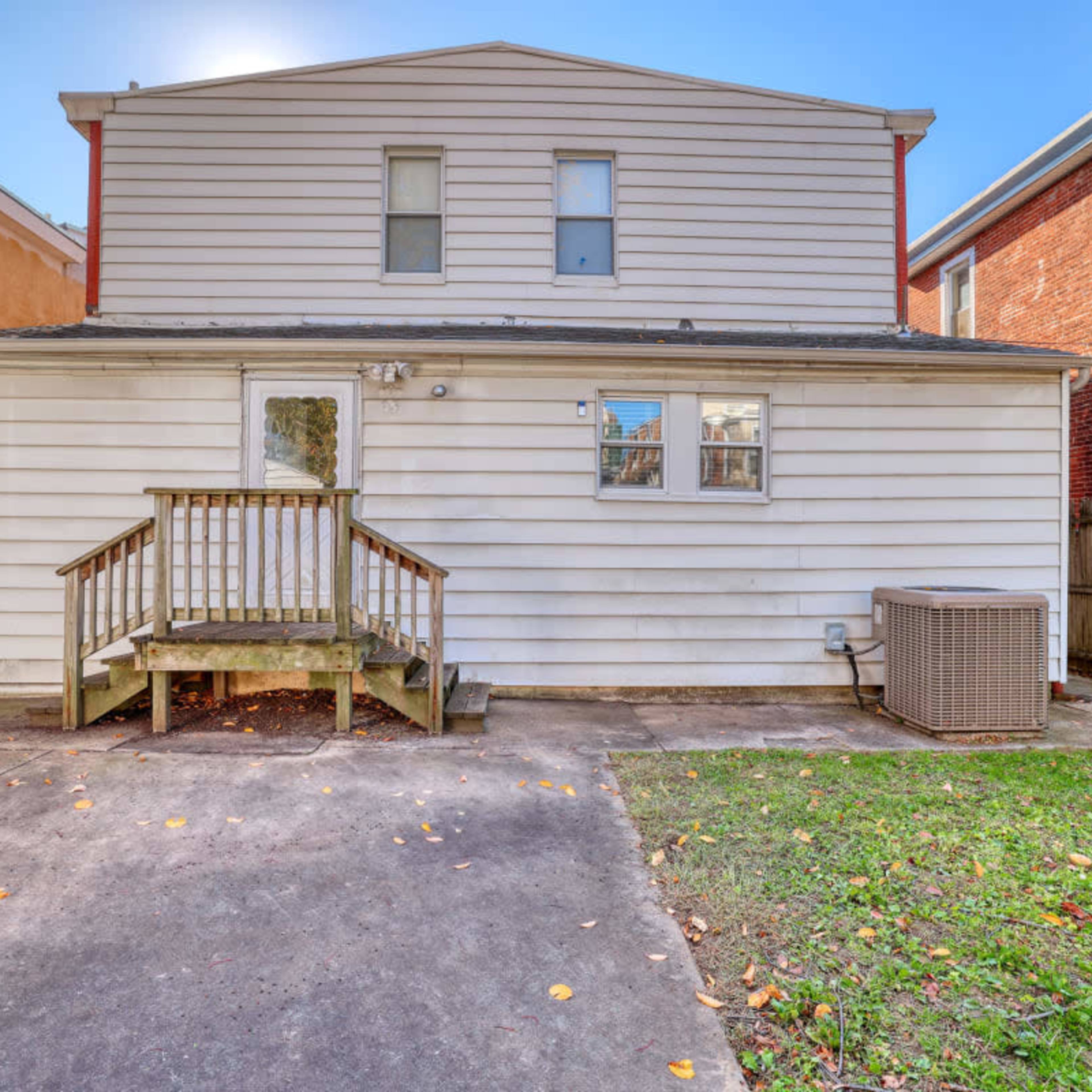 The rear view of a two-story house with a wooden deck, surrounded by a fenced yard and a concrete driveway.