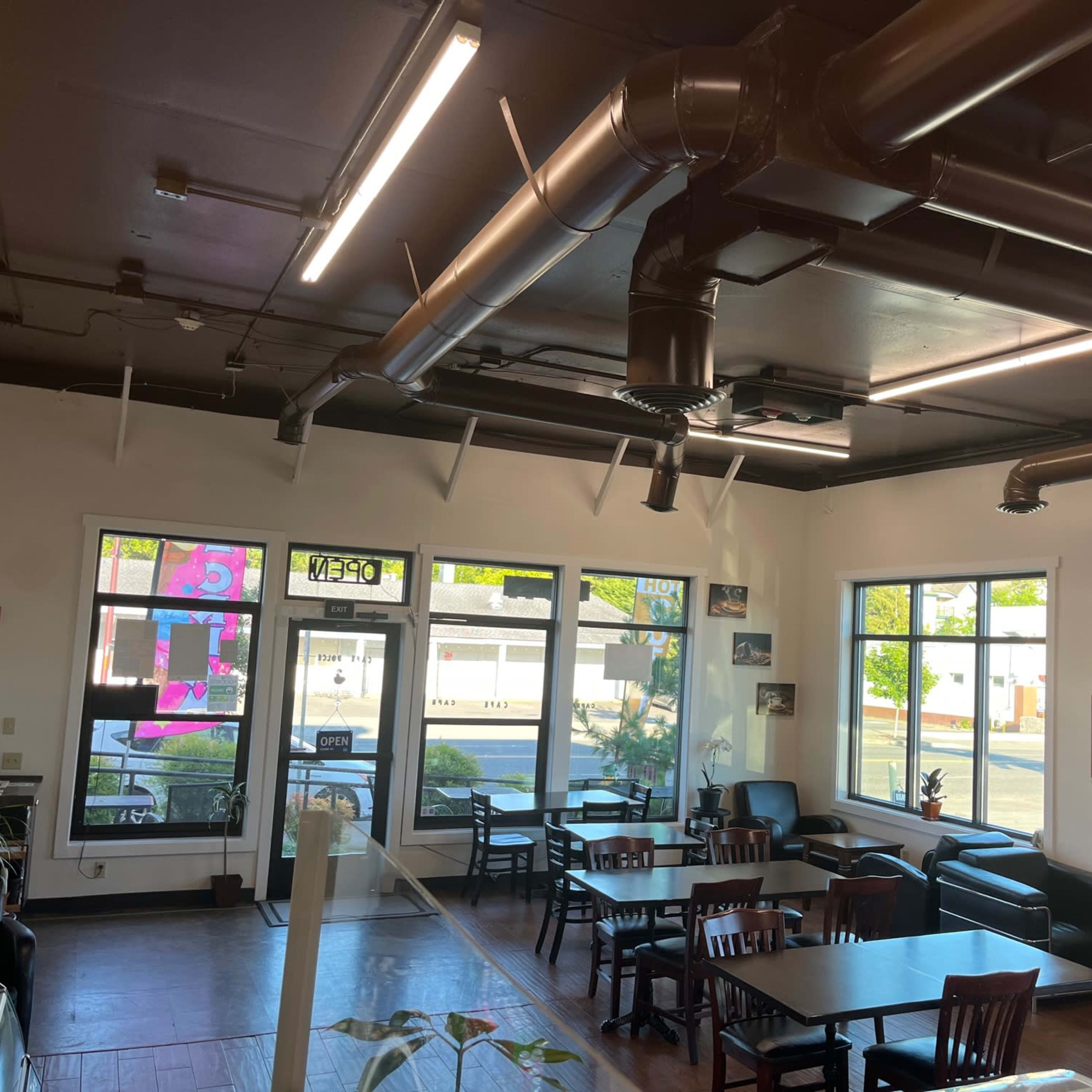 An interior view of a cafe with several tables and chairs, large windows letting in natural light, and exposed ductwork on the ceiling.