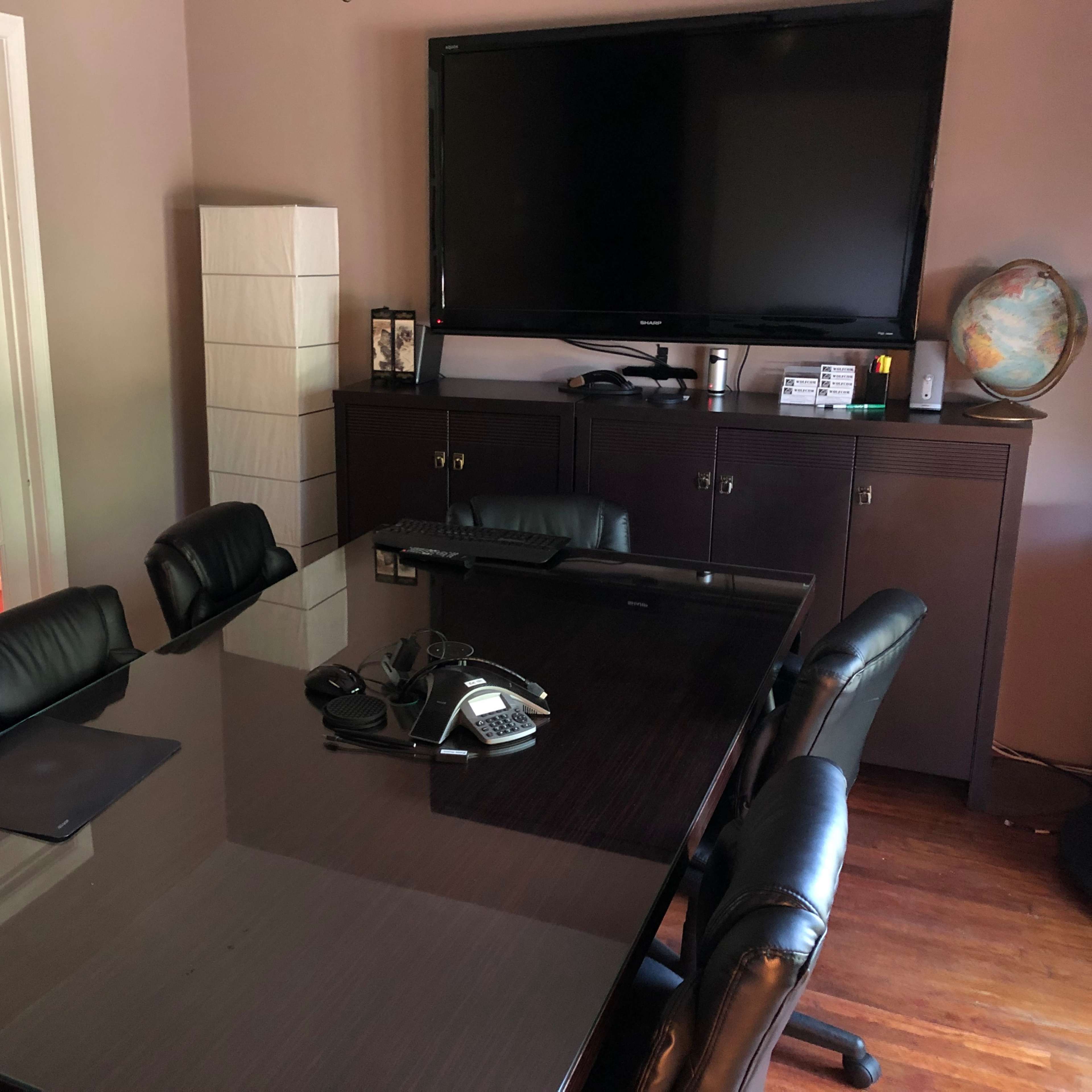 A modern conference room features a large table surrounded by black chairs, with a television mounted on the wall and a phone on the table.