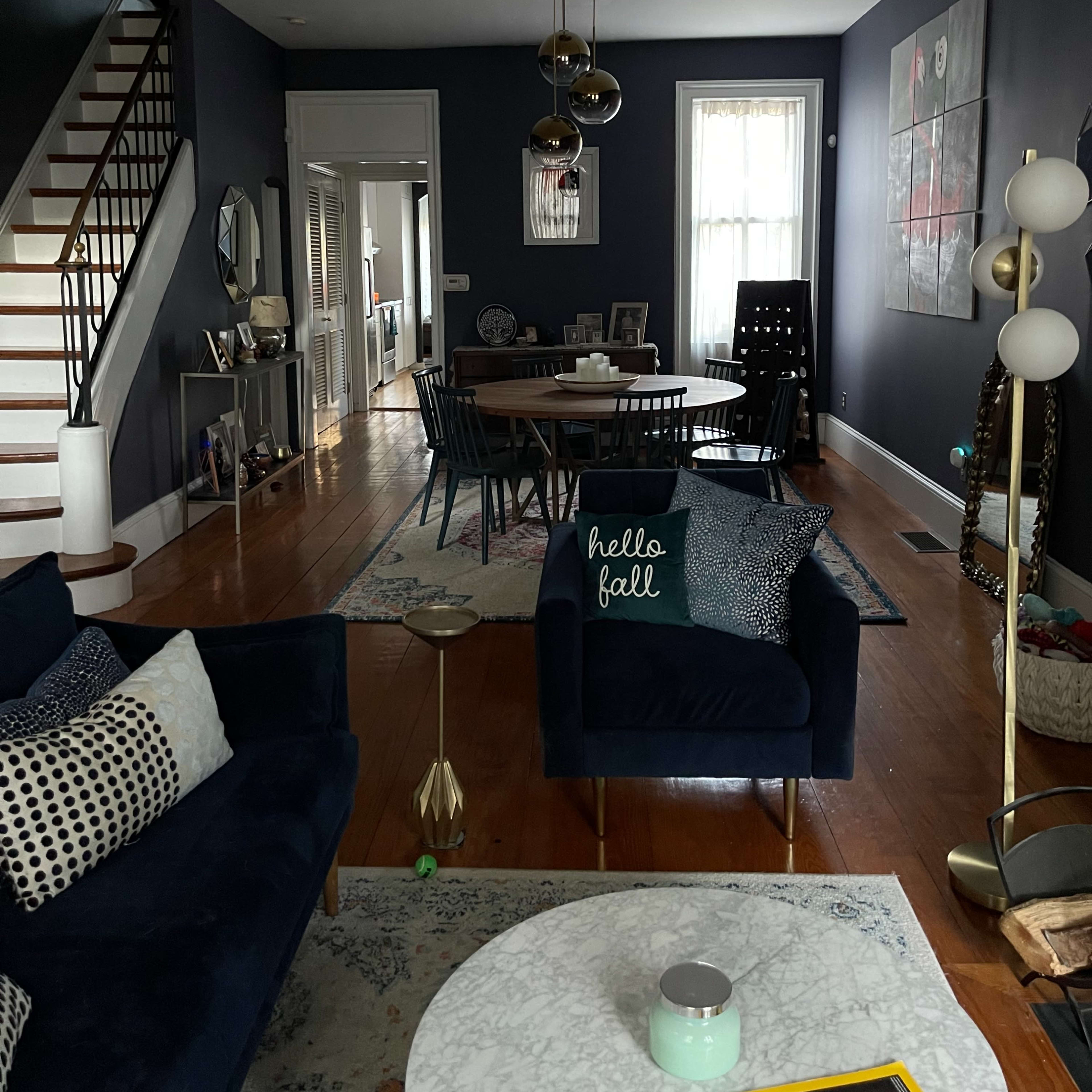 A living room with a dark blue wall, a seating area with a navy couch and patterned cushions, a dining table in the background, and a staircase leading to an upper floor.