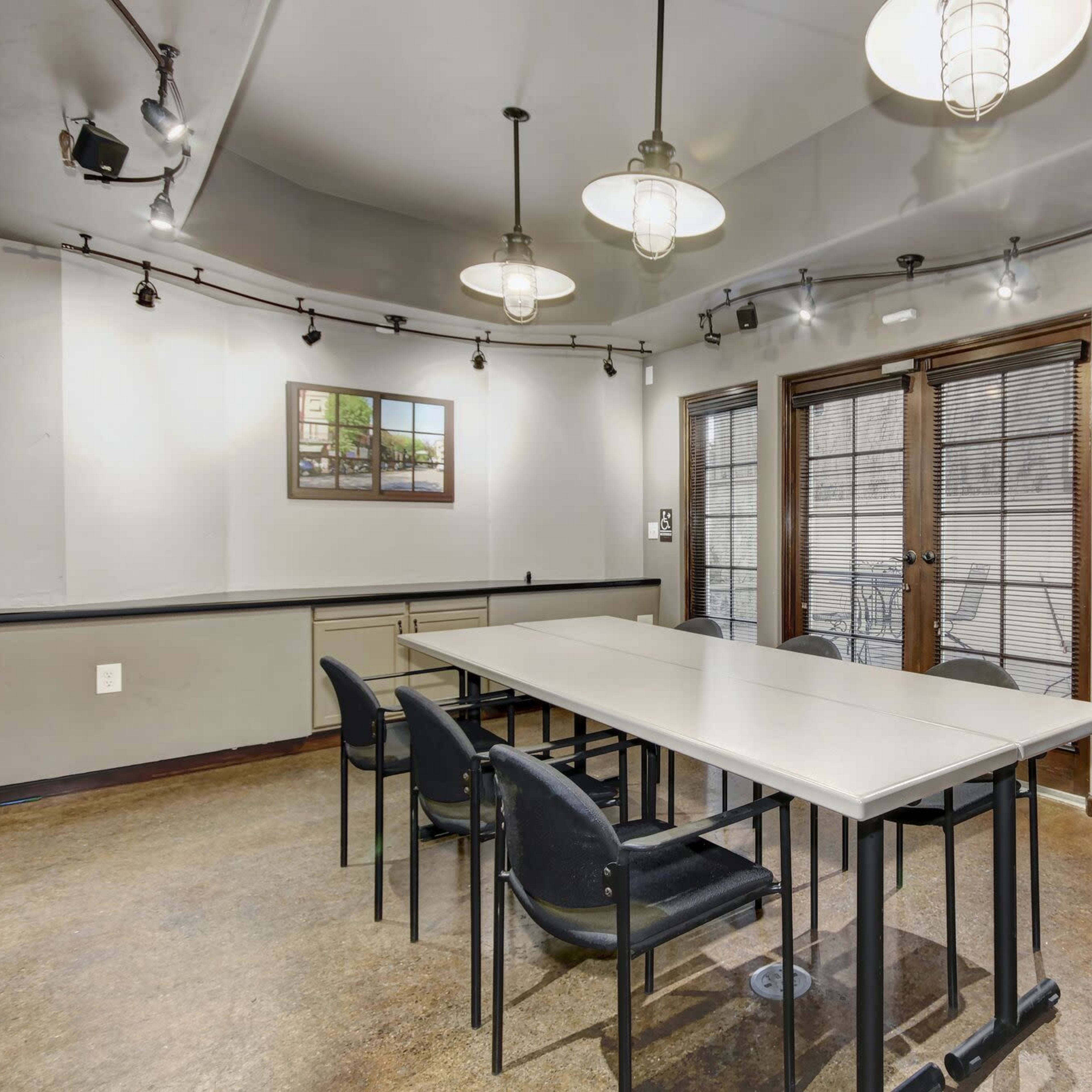 A meeting room with a long table, black chairs, and large windows with blinds, illuminated by pendant lights.