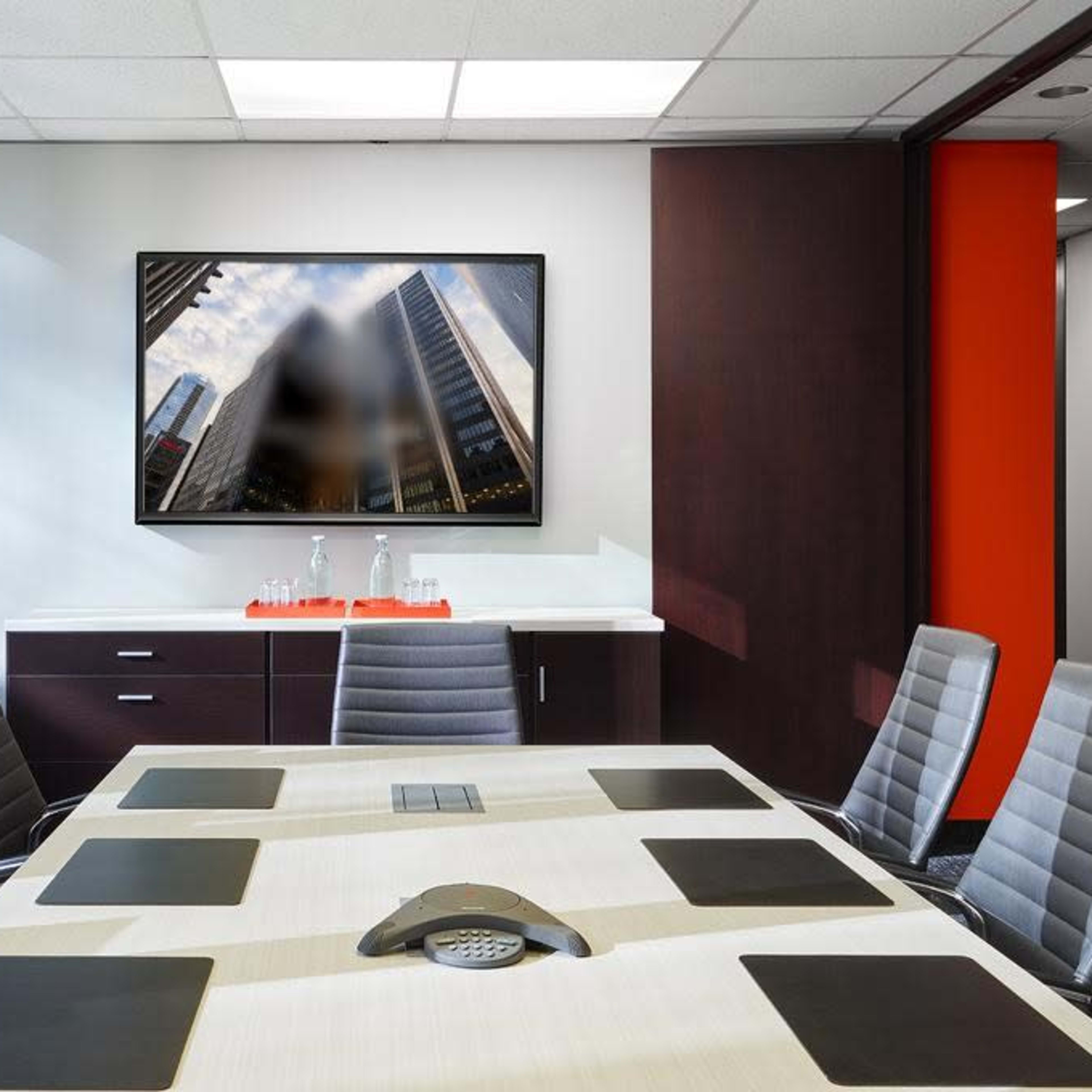 A boardroom features a large table surrounded by chairs and a wall-mounted television displaying a cityscape.