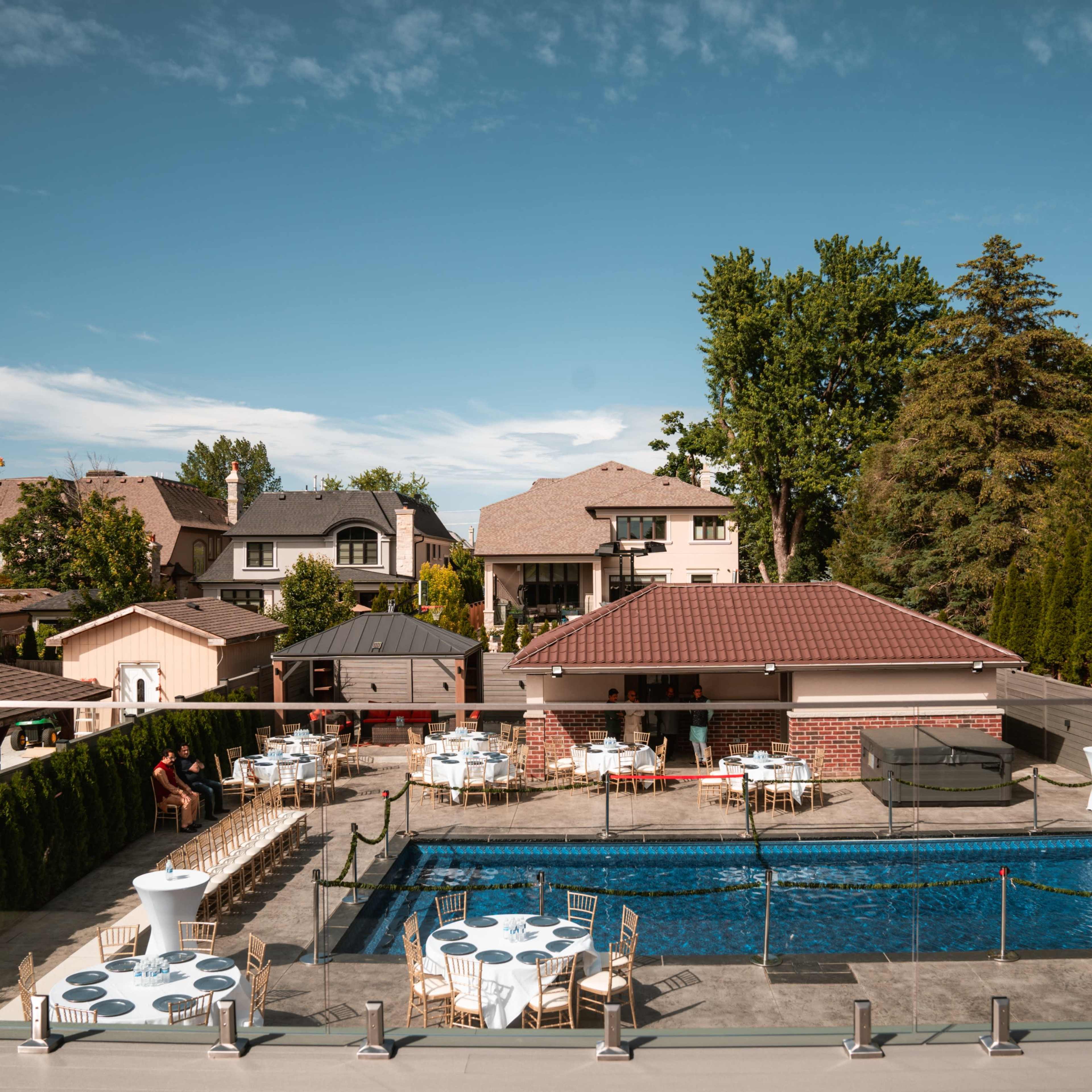 A backyard scene features a swimming pool surrounded by tables and chairs, with houses and trees in the background.