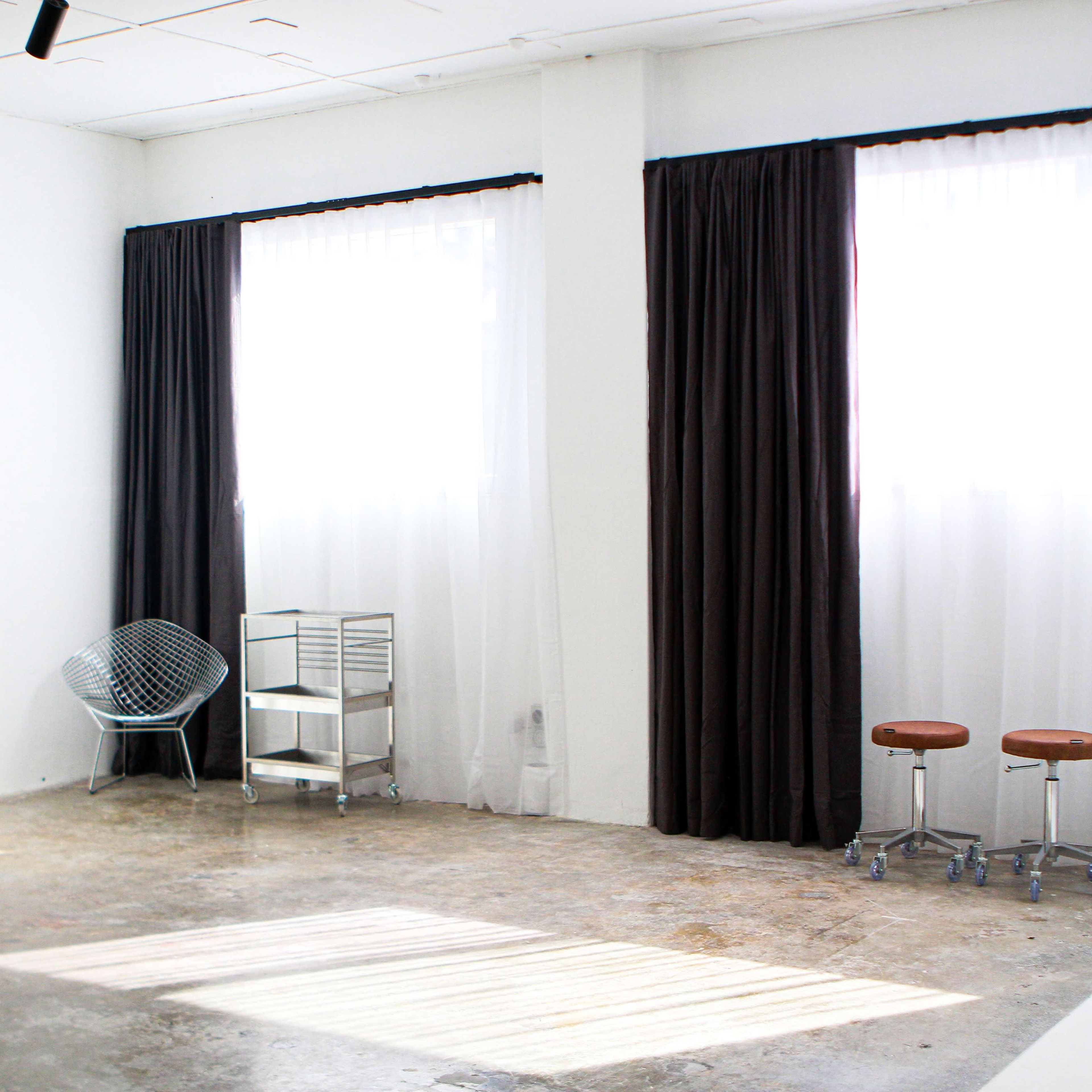 A bright, minimalist room with two large windows, dark curtains, a wire chair, a metal cart, and two stools.