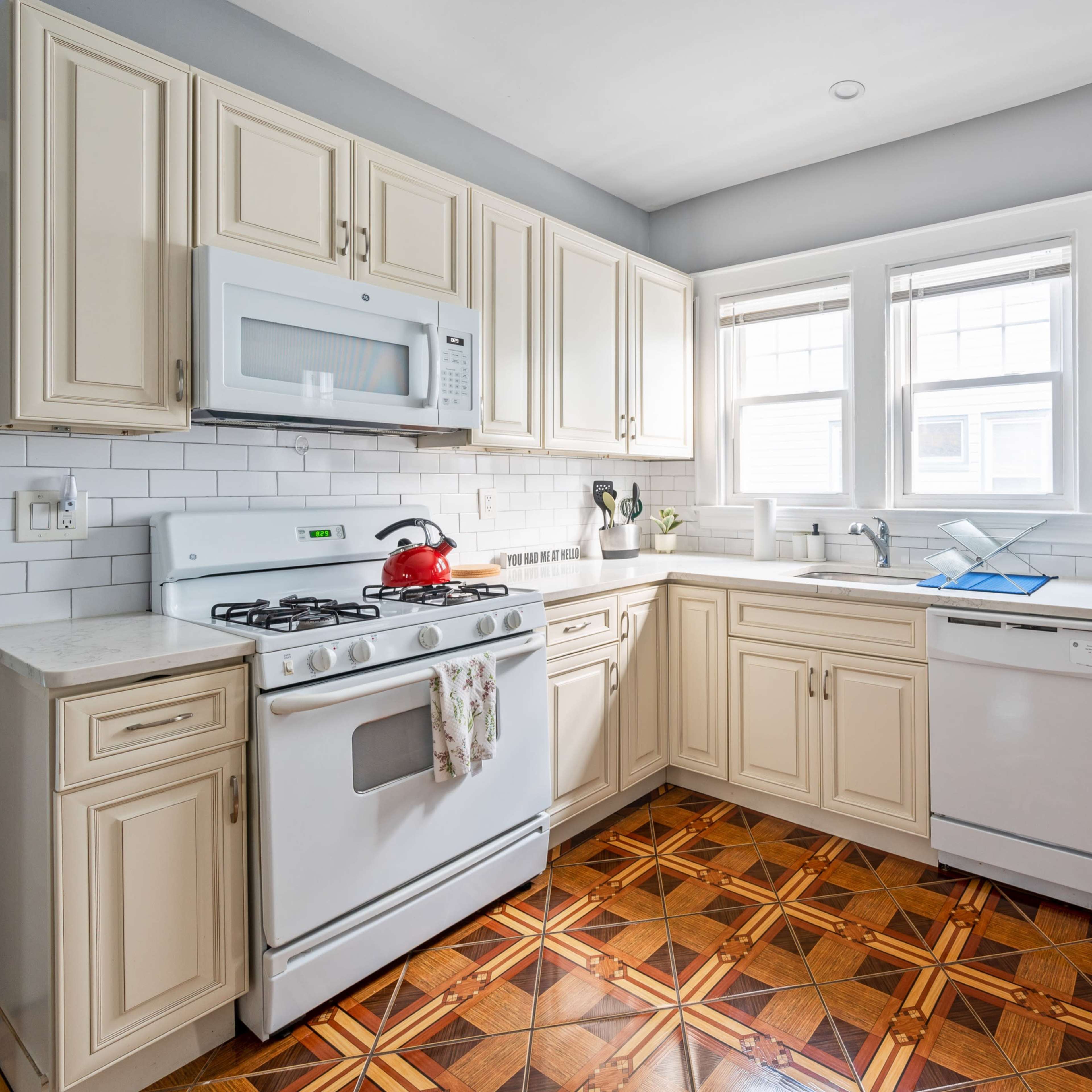 The kitchen features cream-colored cabinets, a gas stove, a microwave, and a dishwasher, with a tiled floor pattern and ample natural light from the windows.