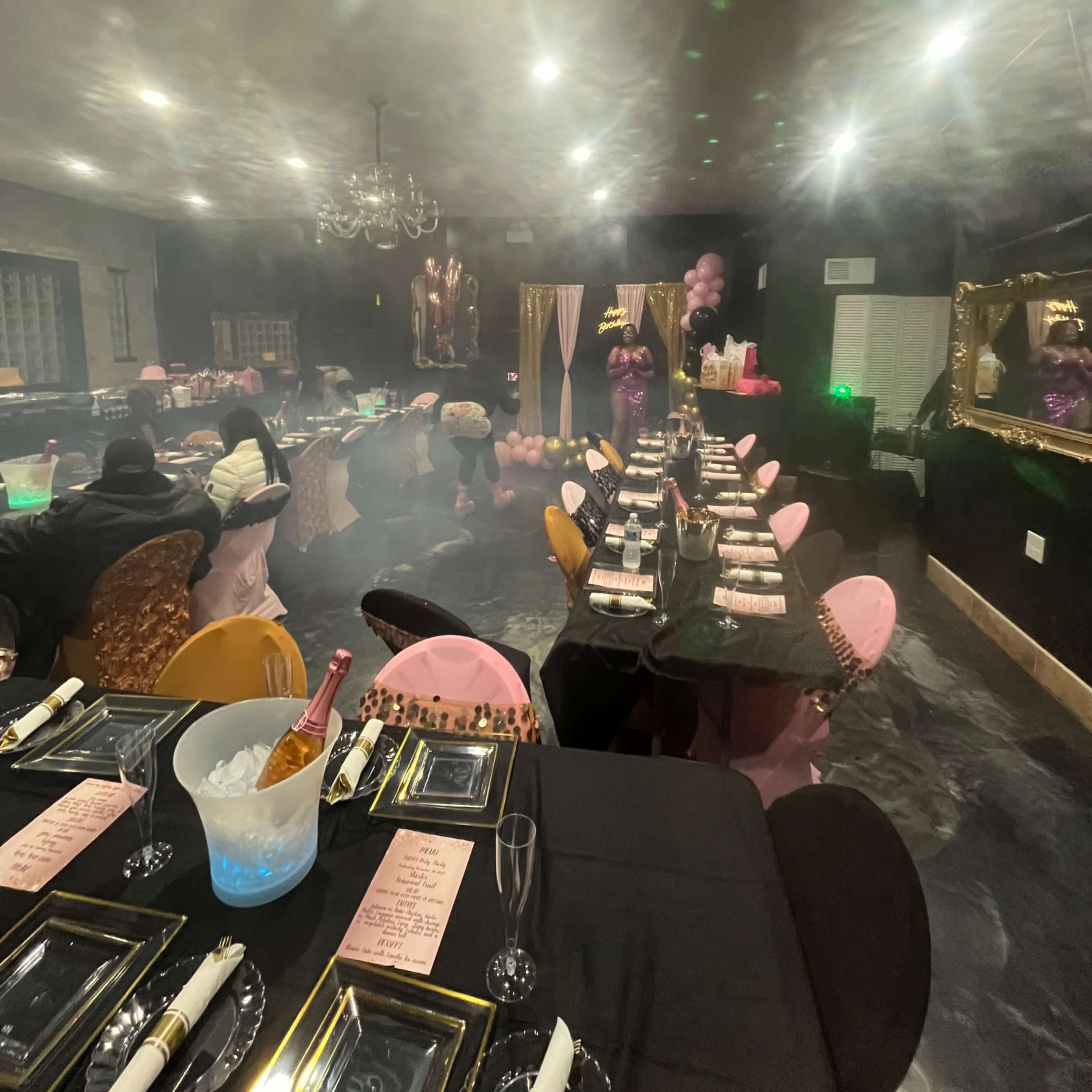 A dimly lit dining room set up for a party, featuring elegantly arranged tables with black tablecloths, colorful chairs, and decorations including balloons and soft lighting.
