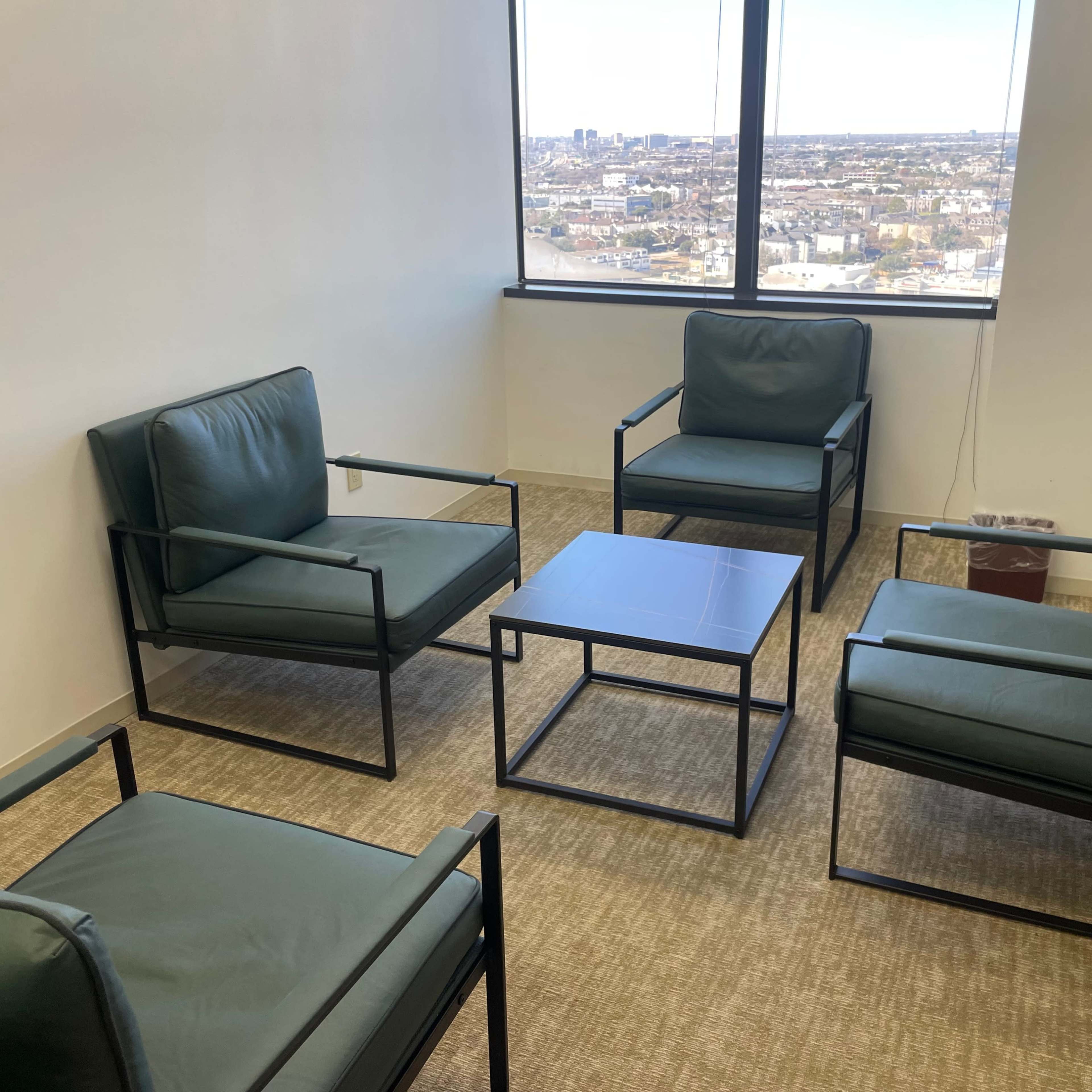 A small meeting room with four green chairs and a square table, overlooking a city skyline through a large window.