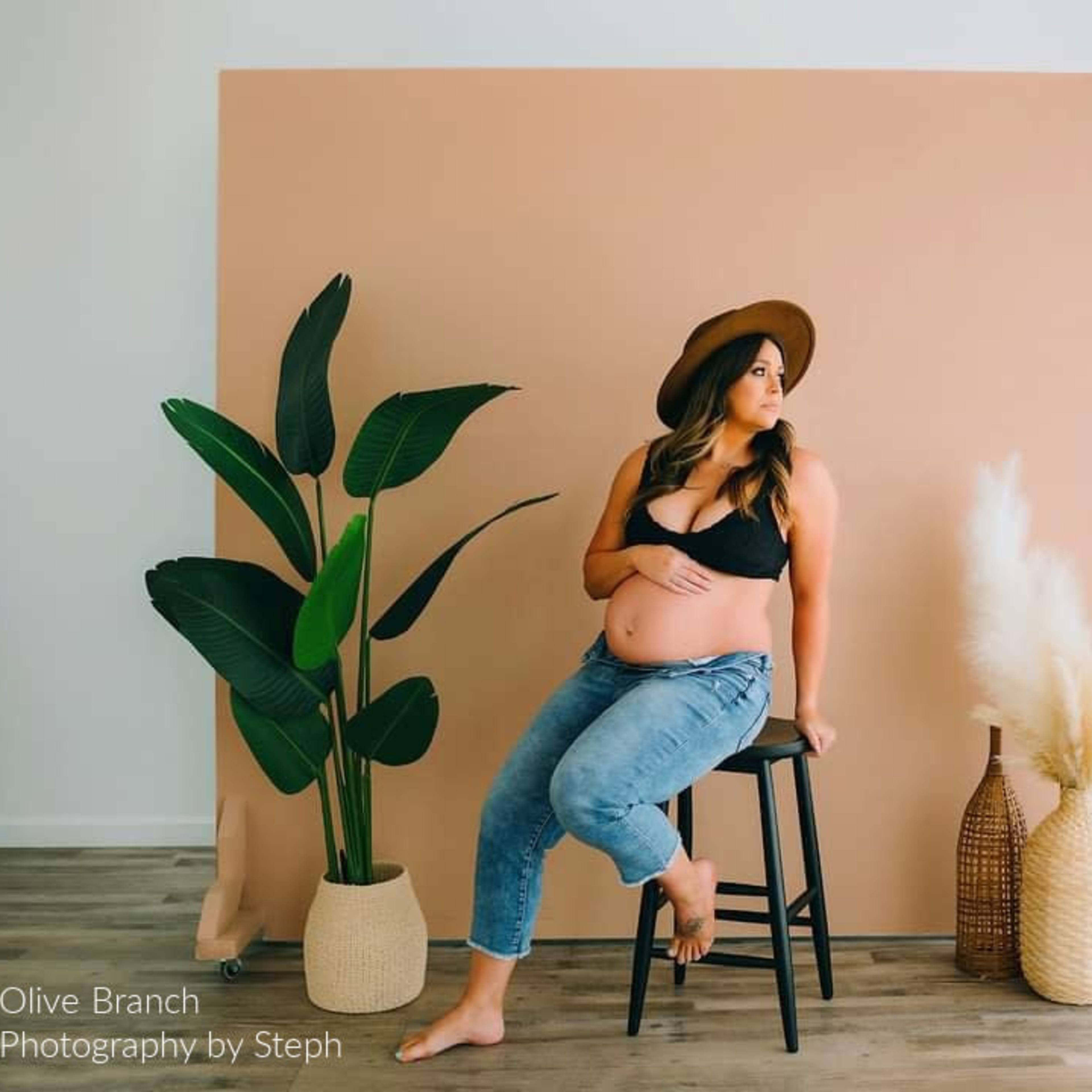 A pregnant woman in a black top and jeans sits on a stool beside a potted plant and decorative vases against a peach-colored backdrop.
