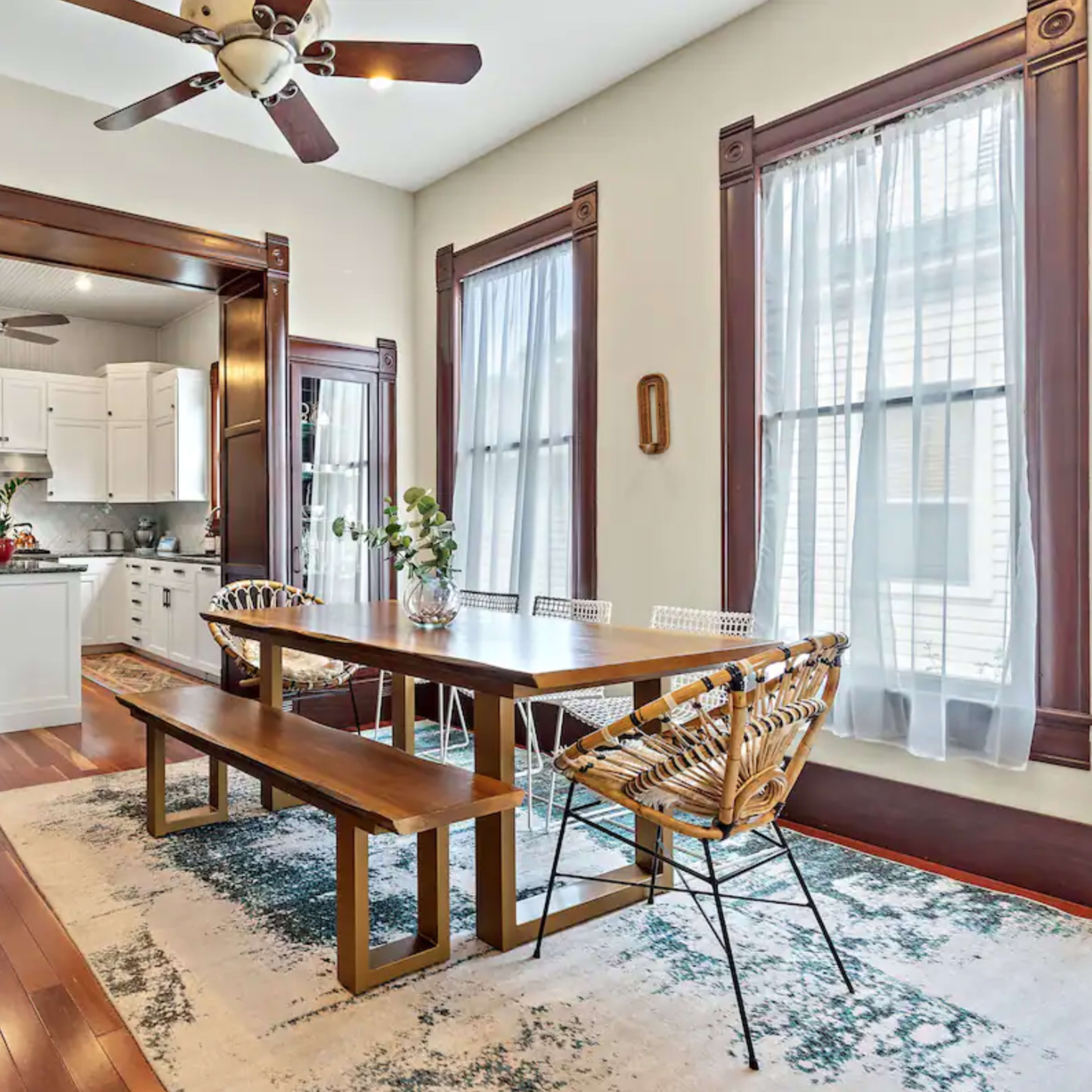 A dining area with a wooden table and benches is adjacent to a kitchen, featuring white cabinetry and large windows, all framed by dark wood trim.