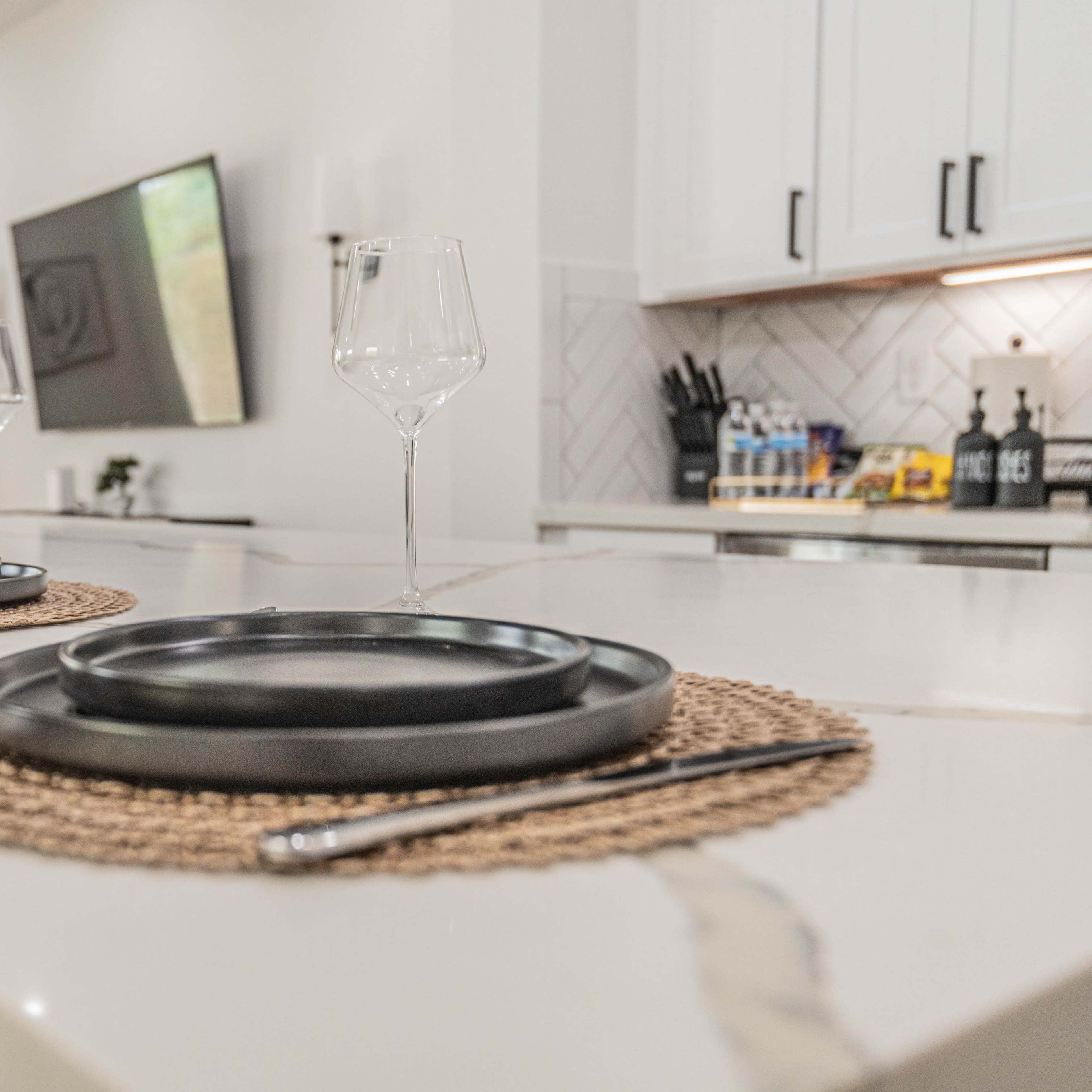 A modern kitchen features a dining island set with black plates and wine glasses, accompanied by woven placemats.