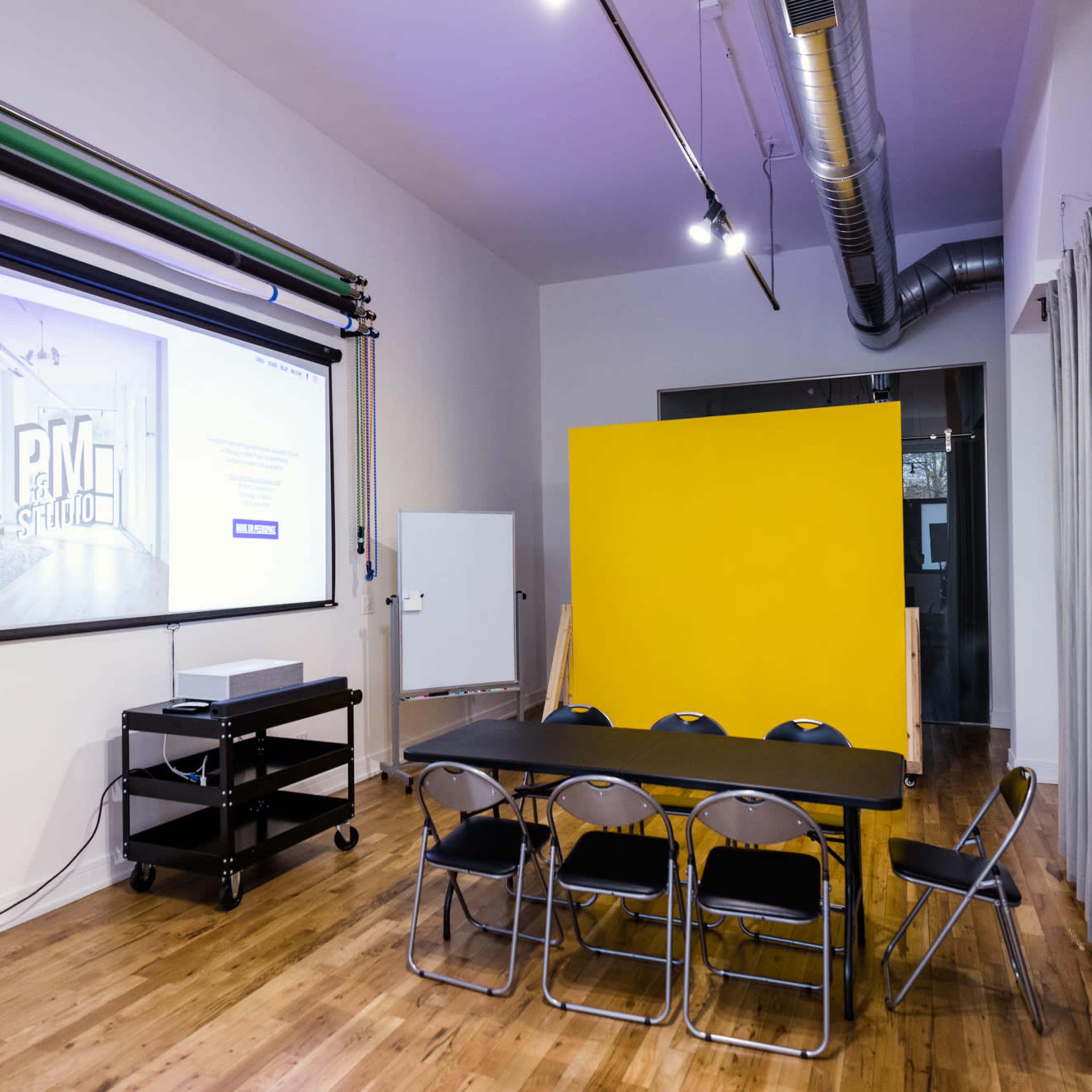 A modern meeting room with a large screen displaying a website, a rectangular table surrounded by chairs, and a bright yellow wall section.