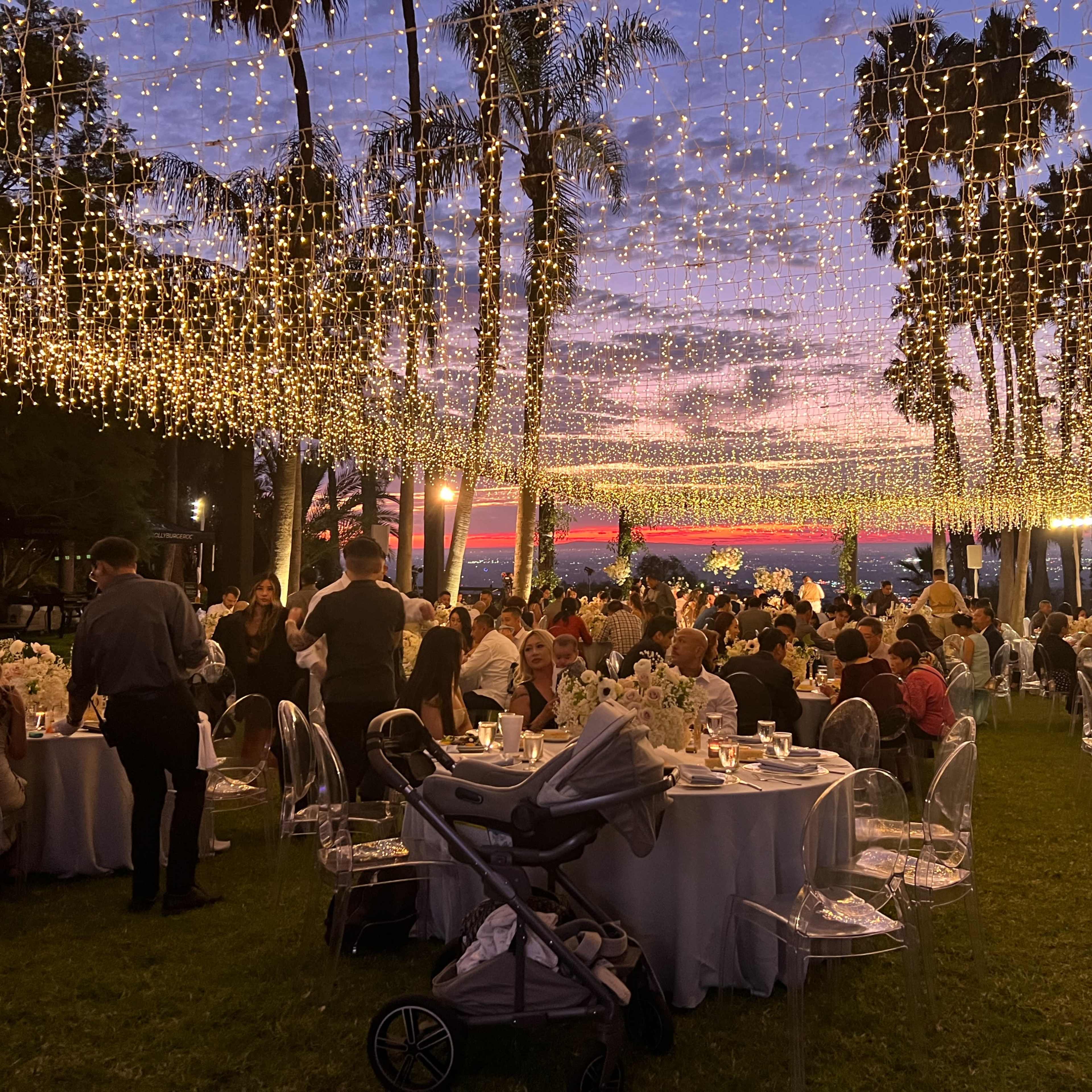A large outdoor event is set up with elegantly decorated tables under string lights, surrounded by palm trees and guests enjoying a sunset view.