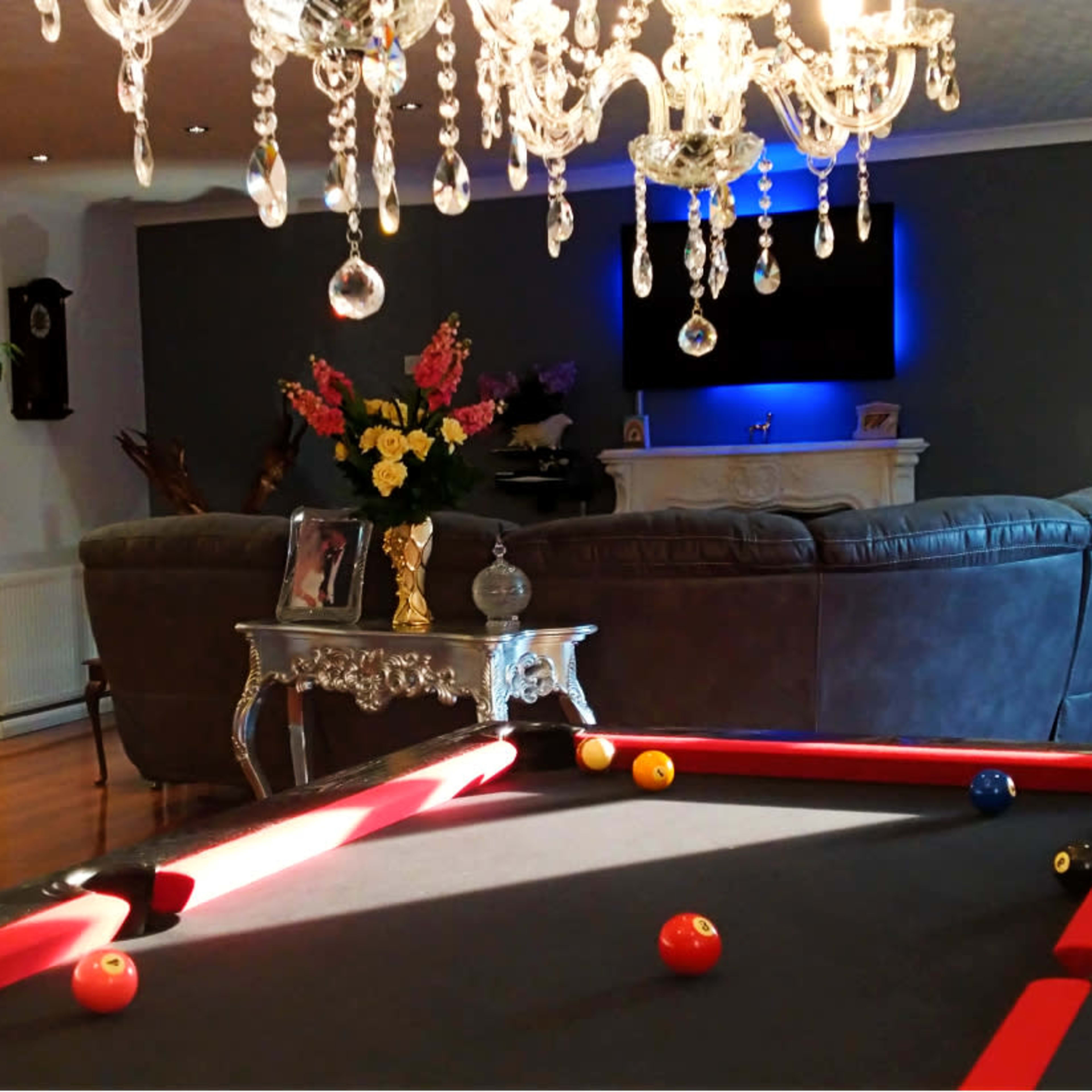 A billiards table in the foreground with a living room featuring a sofa, a fireplace, and decorative plants in the background.