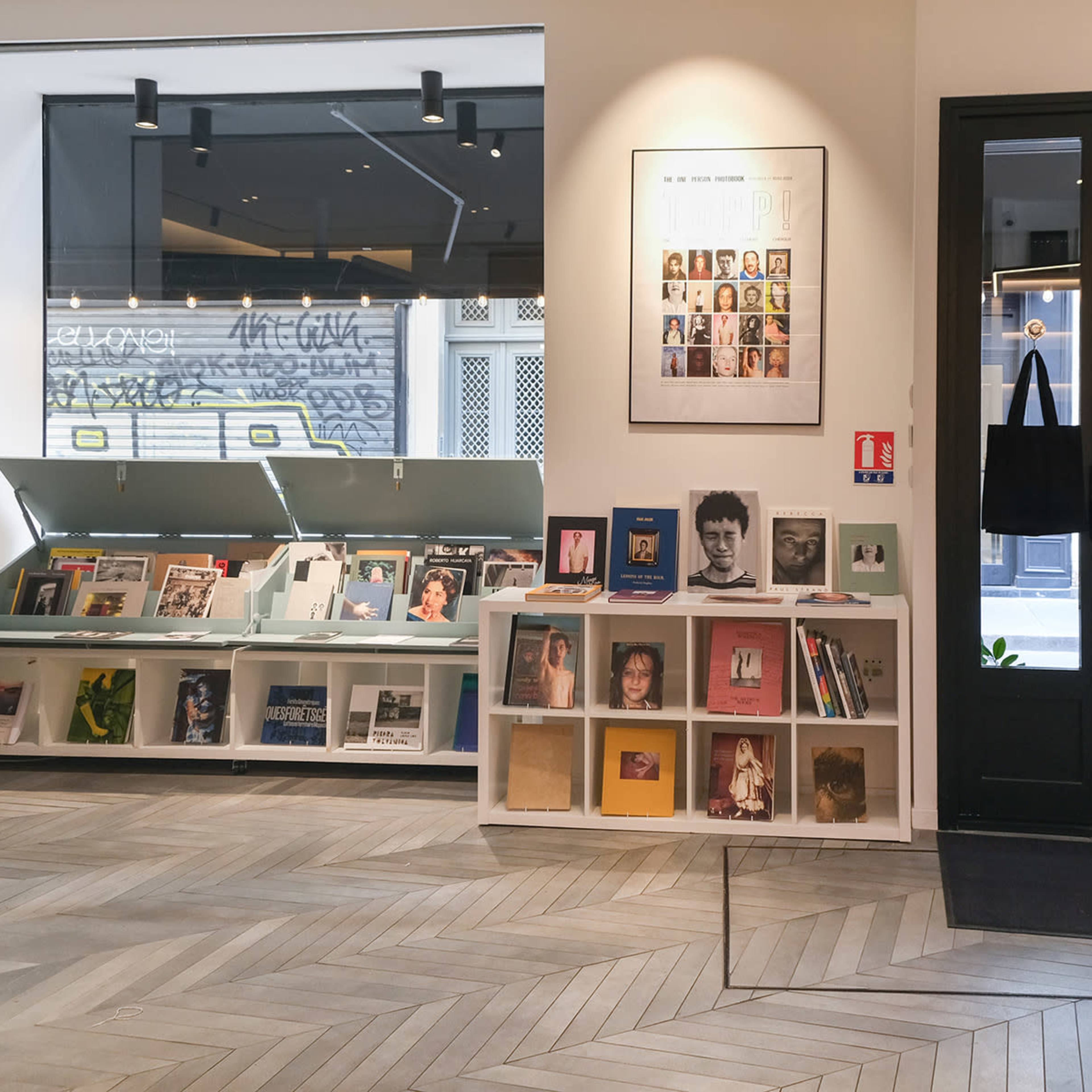 A modern interior of a bookstore or gallery featuring shelves of books and photographs on the left, and a glass entrance door on the right.