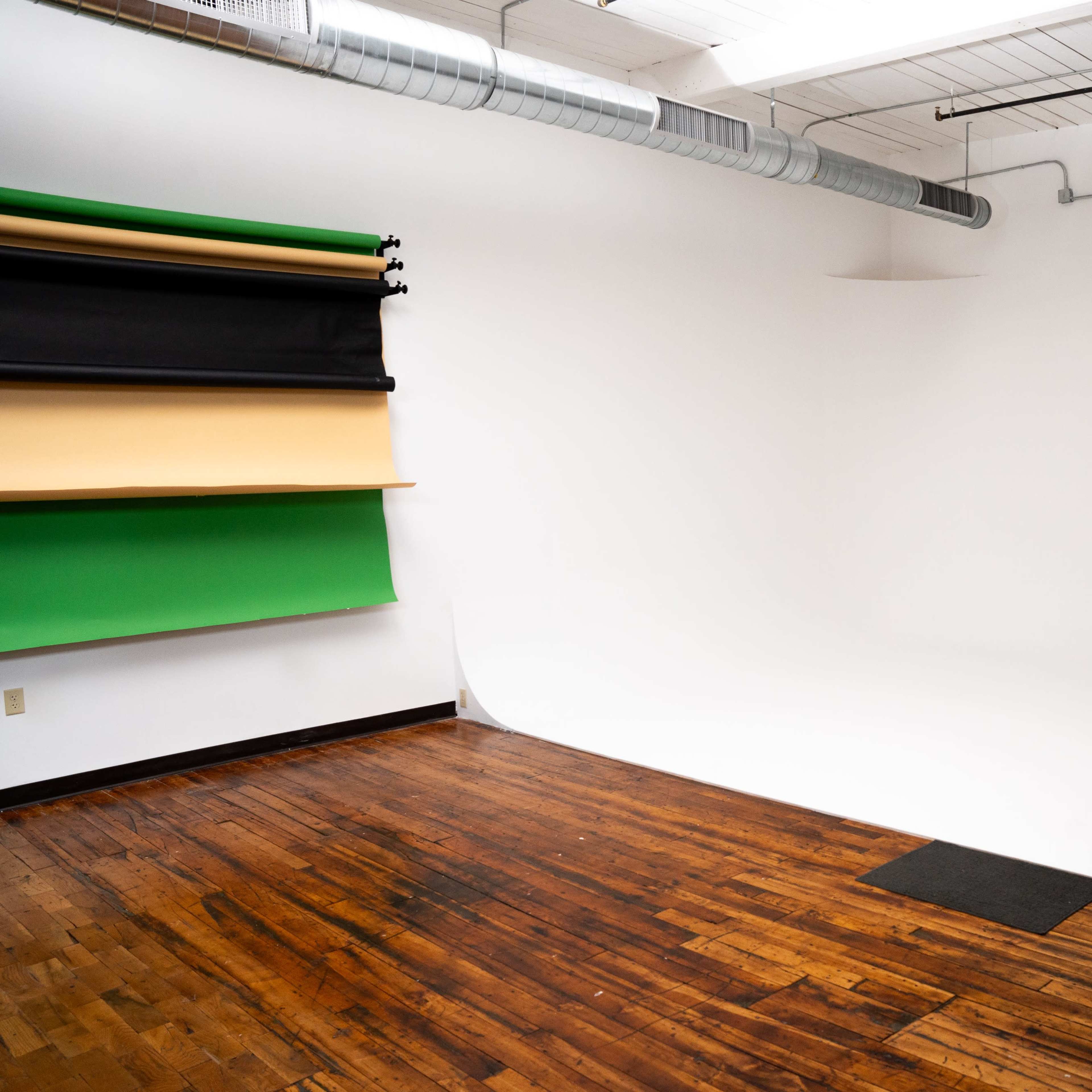 A spacious studio with a seamless backdrop of white and colored backdrops hanging on the wall, along with wooden flooring.