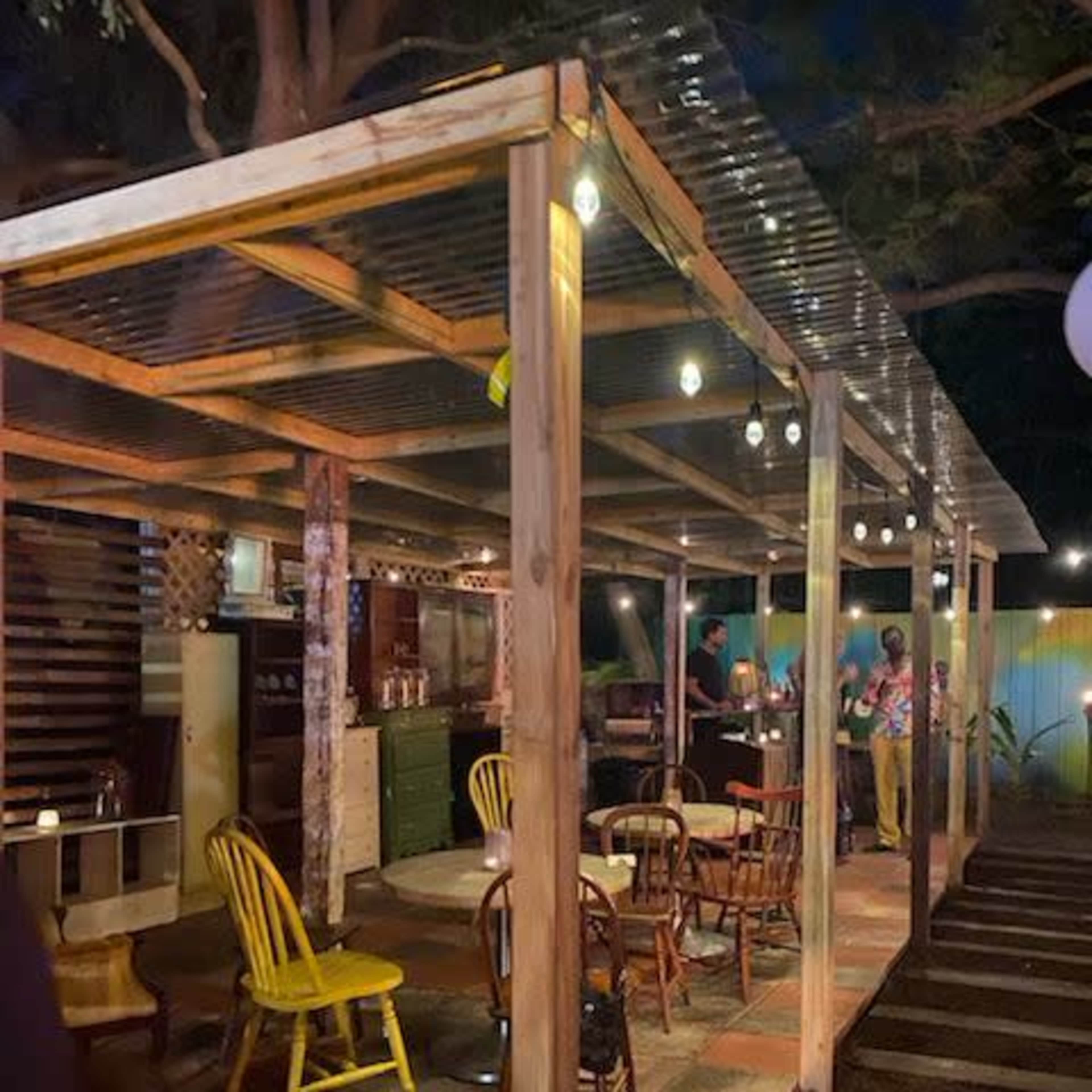 A rustic outdoor dining area with wooden chairs and tables under a transparent roof, illuminated by string lights in a nighttime setting.