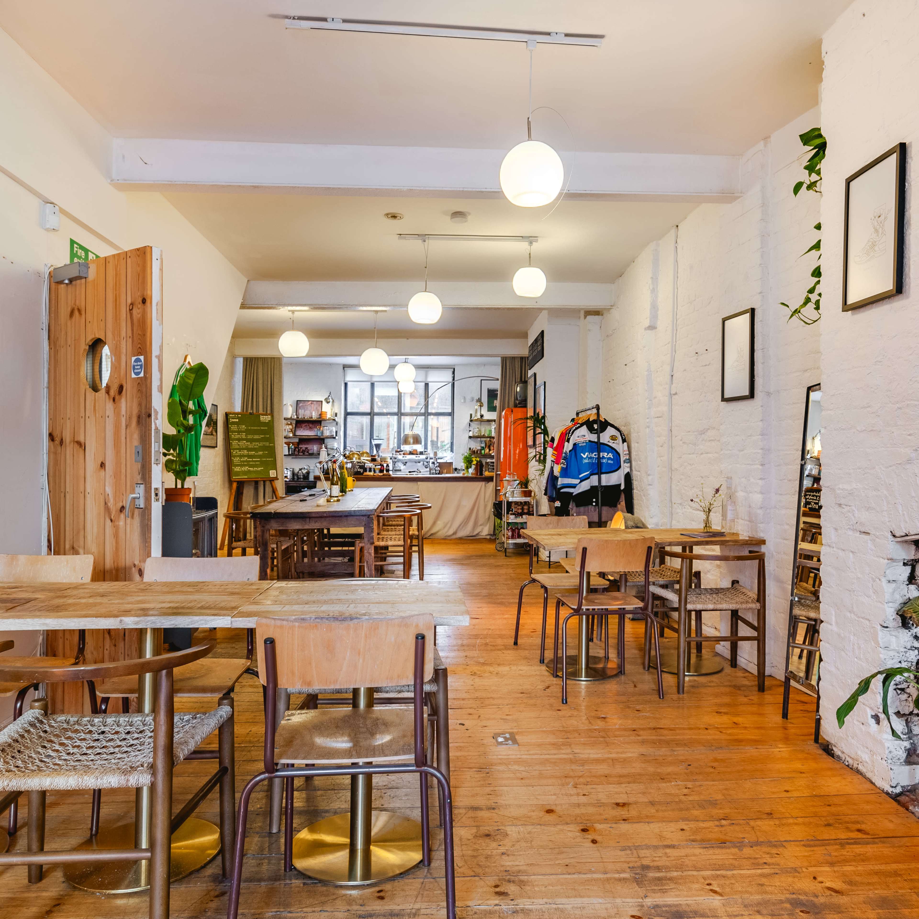 An open, well-lit space with wooden floors, featuring several tables and chairs, a fireplace, and shelves with various items in the background.