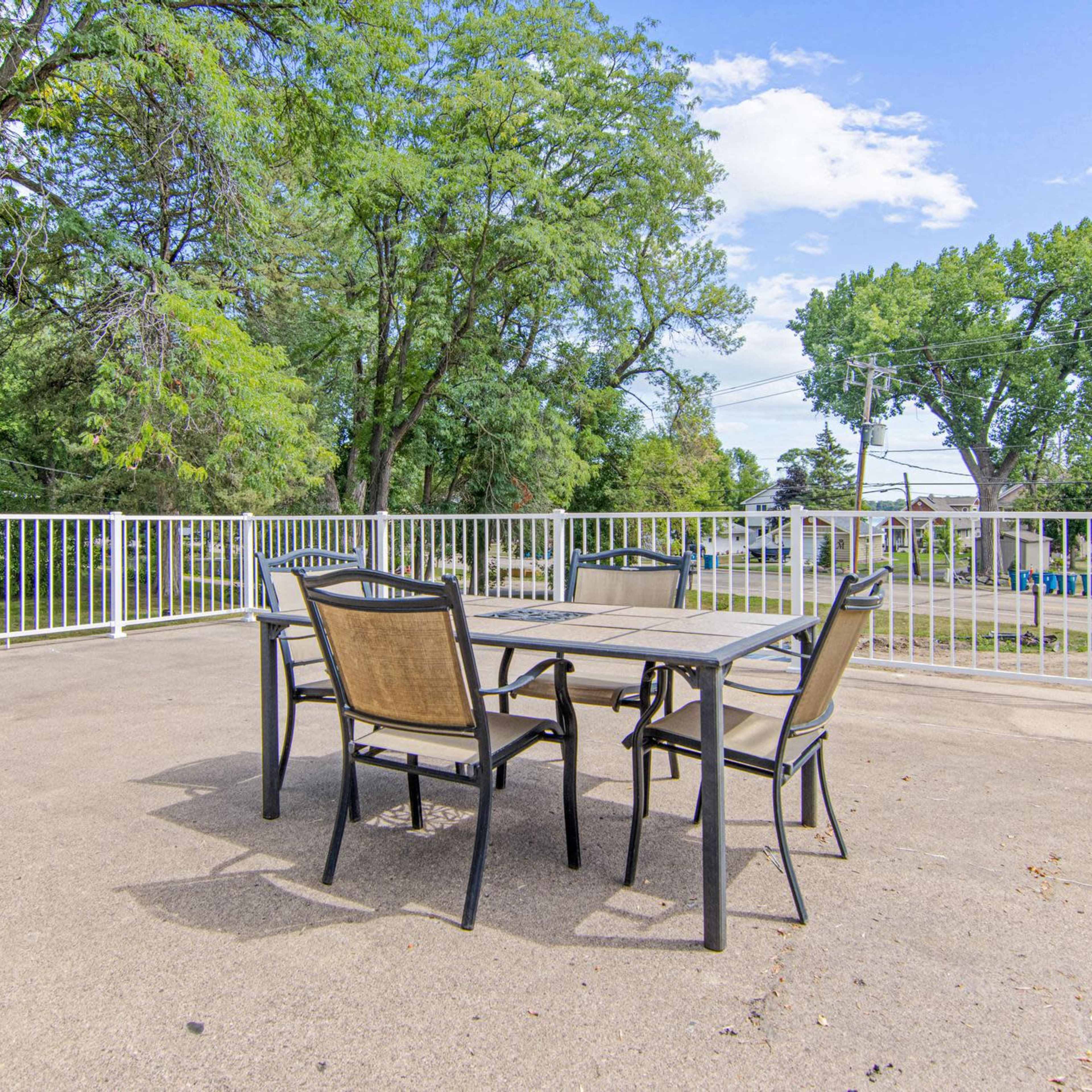 A patio area features a table and chairs surrounded by a white railing and green trees in the background.