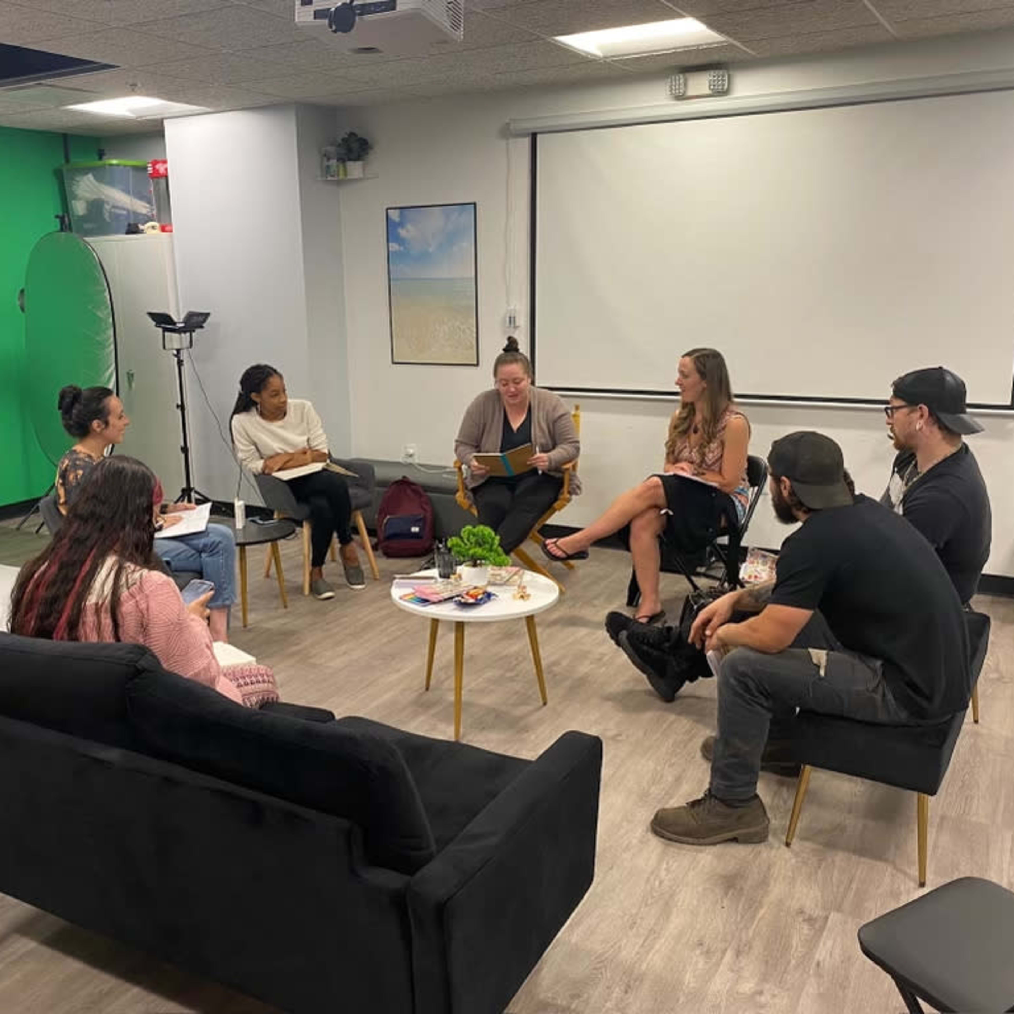 A group of six individuals sits in a modern meeting room, engaged in discussion around a coffee table.