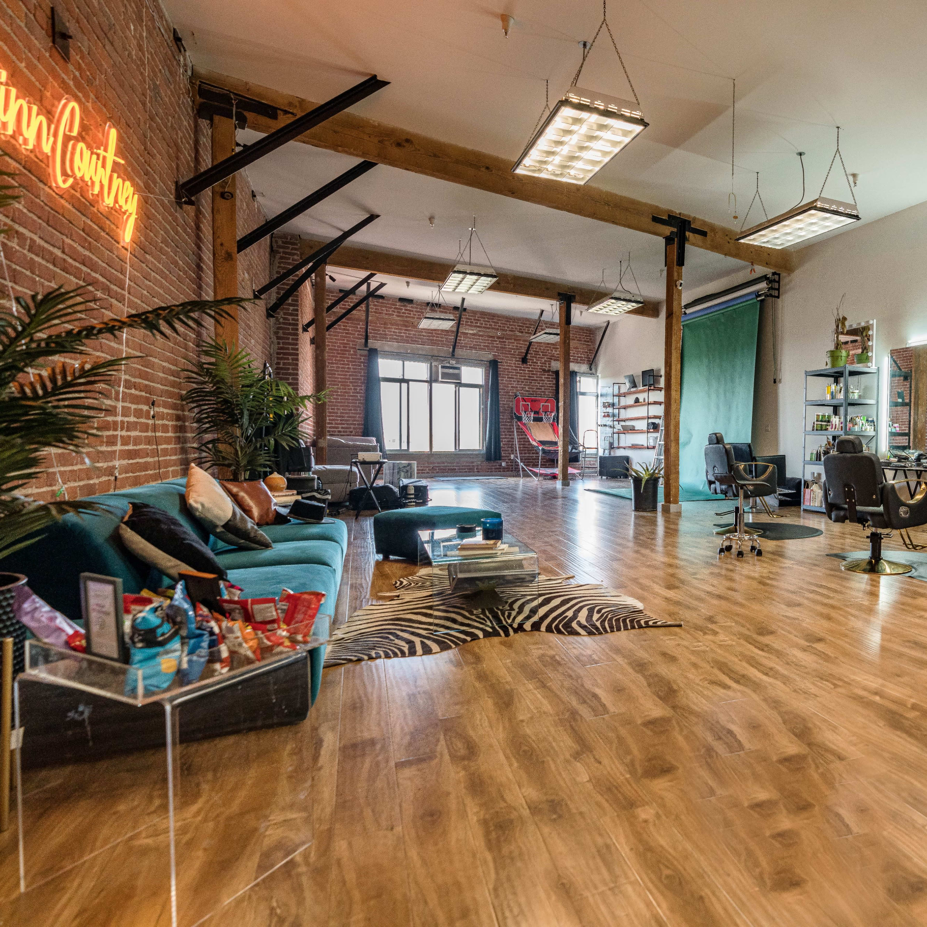 A spacious, modern studio with wooden flooring, large windows, and a mix of seating areas, including a teal sofa and several salon-style chairs.