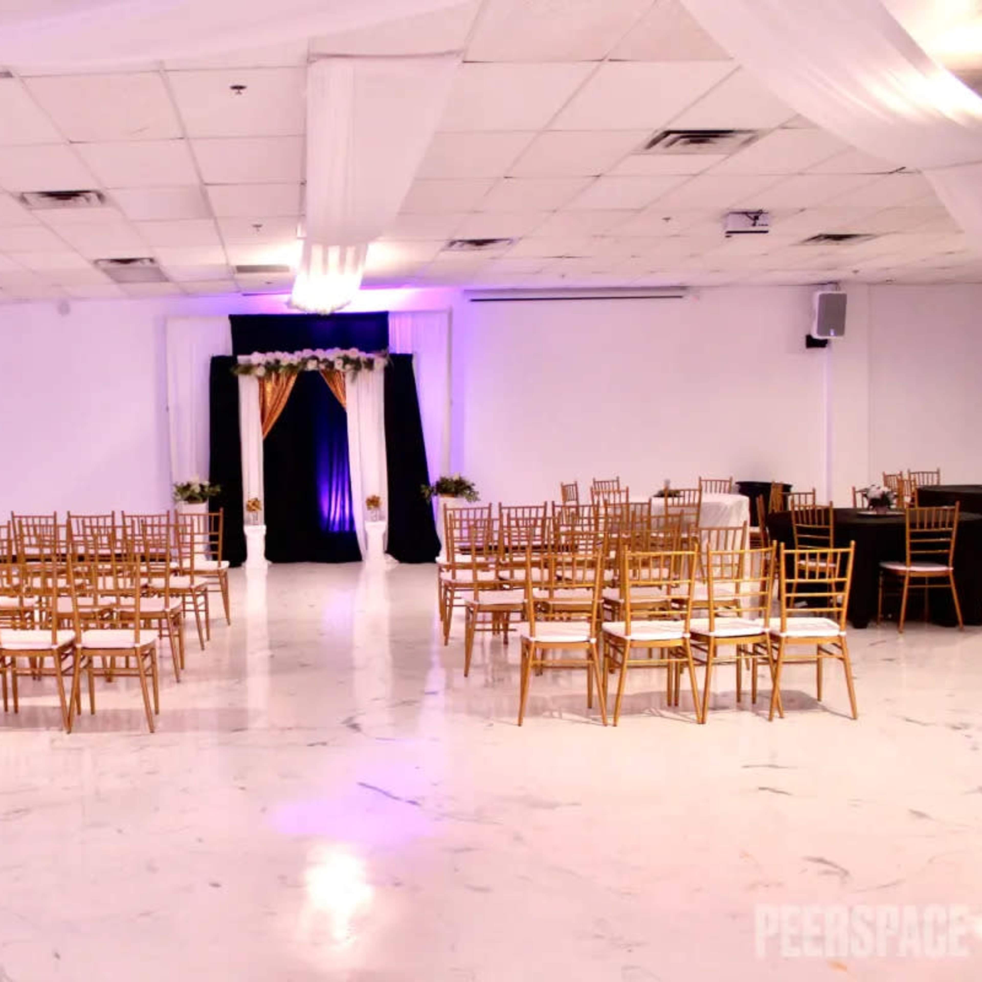 An event space arranged with gold chairs and tables set for a gathering, featuring a decorative backdrop at one end.
