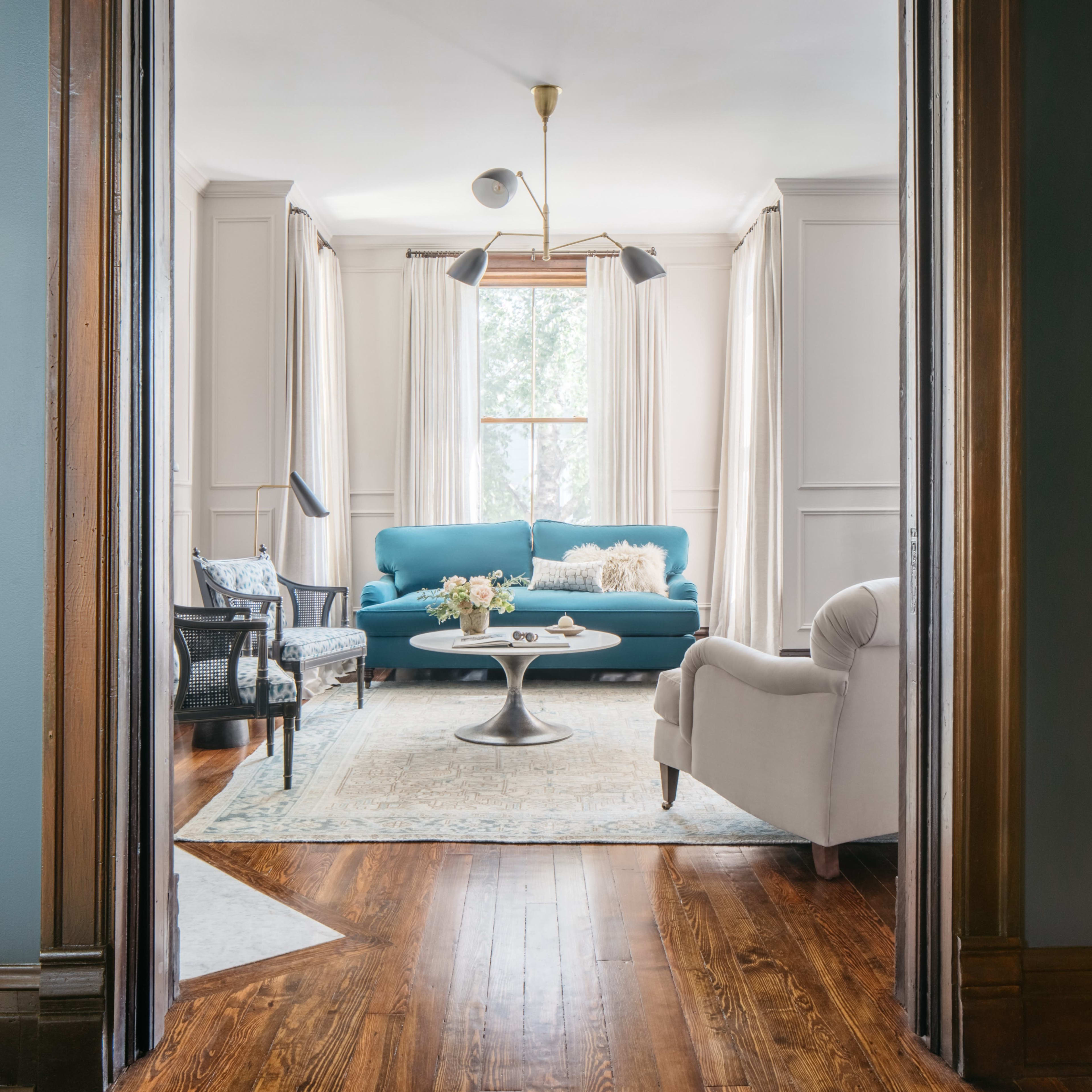 A bright living room features a blue sofa, a circular coffee table, and large windows with sheer curtains.