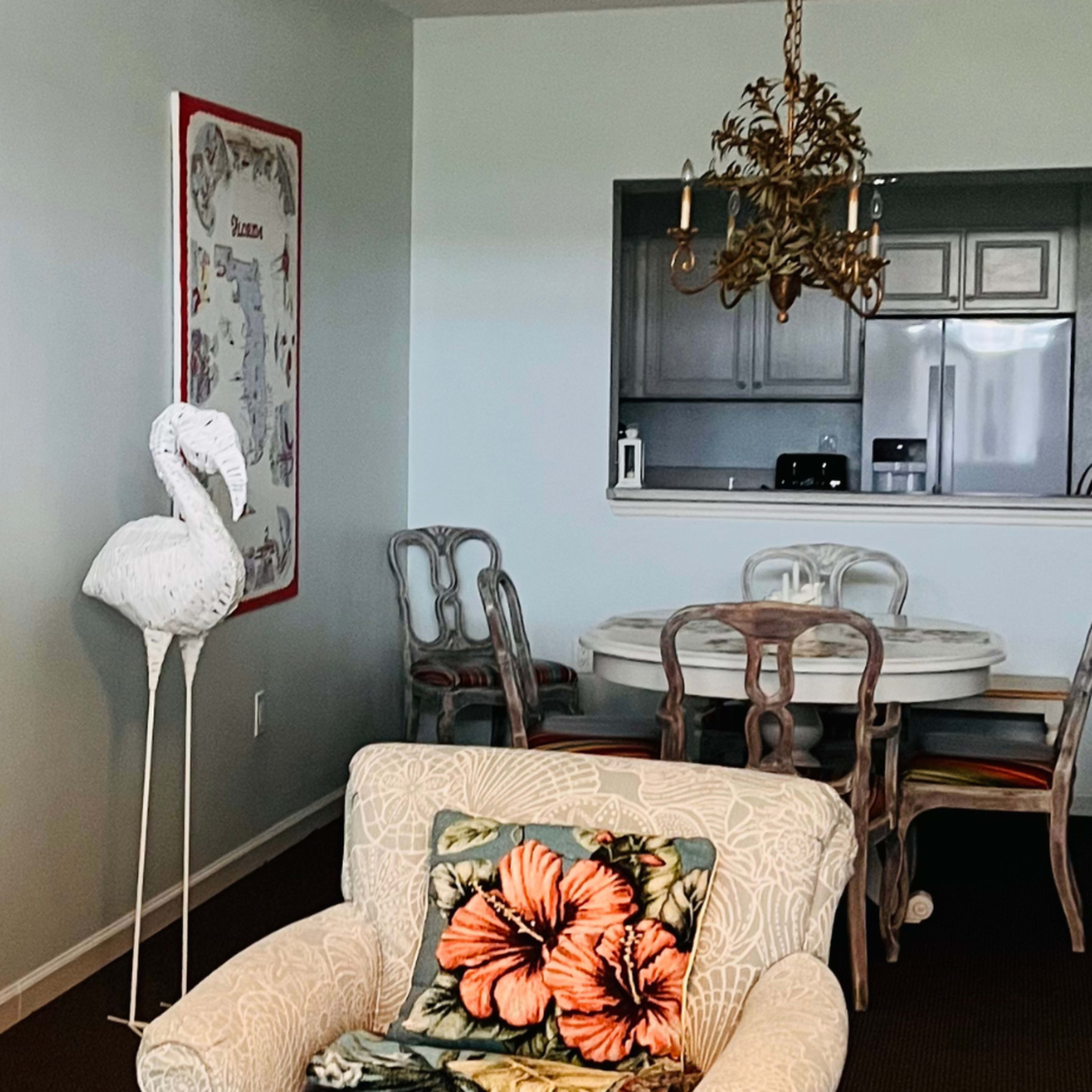 A light-colored living room featuring a patterned armchair, a lamp, a flamingo decoration, and a dining area in the background with a round table and chairs.