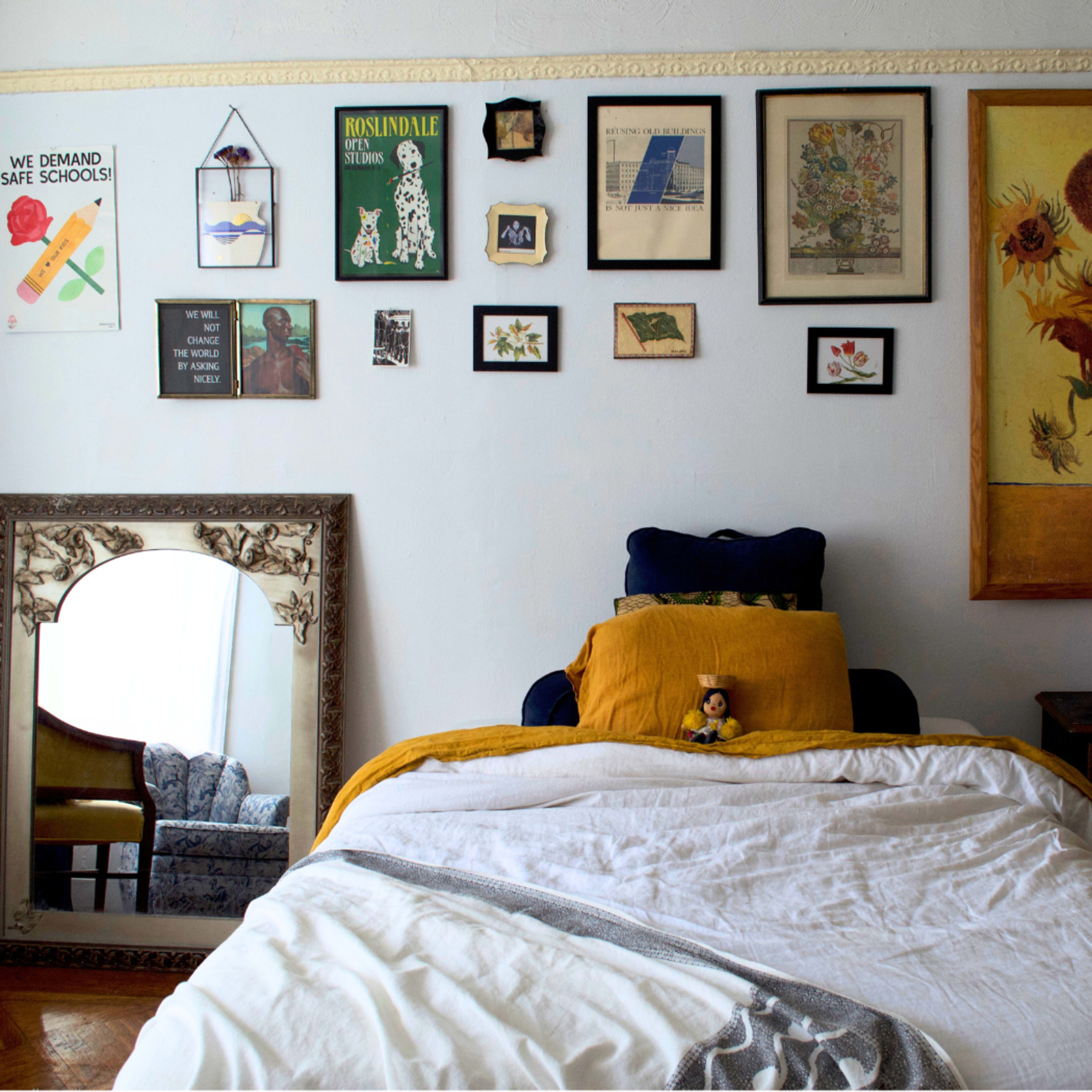 A bedroom features a bed with a white comforter, a blue pillow, and a yellow throw, surrounded by an eclectic display of framed artwork and a large mirror on the wall.