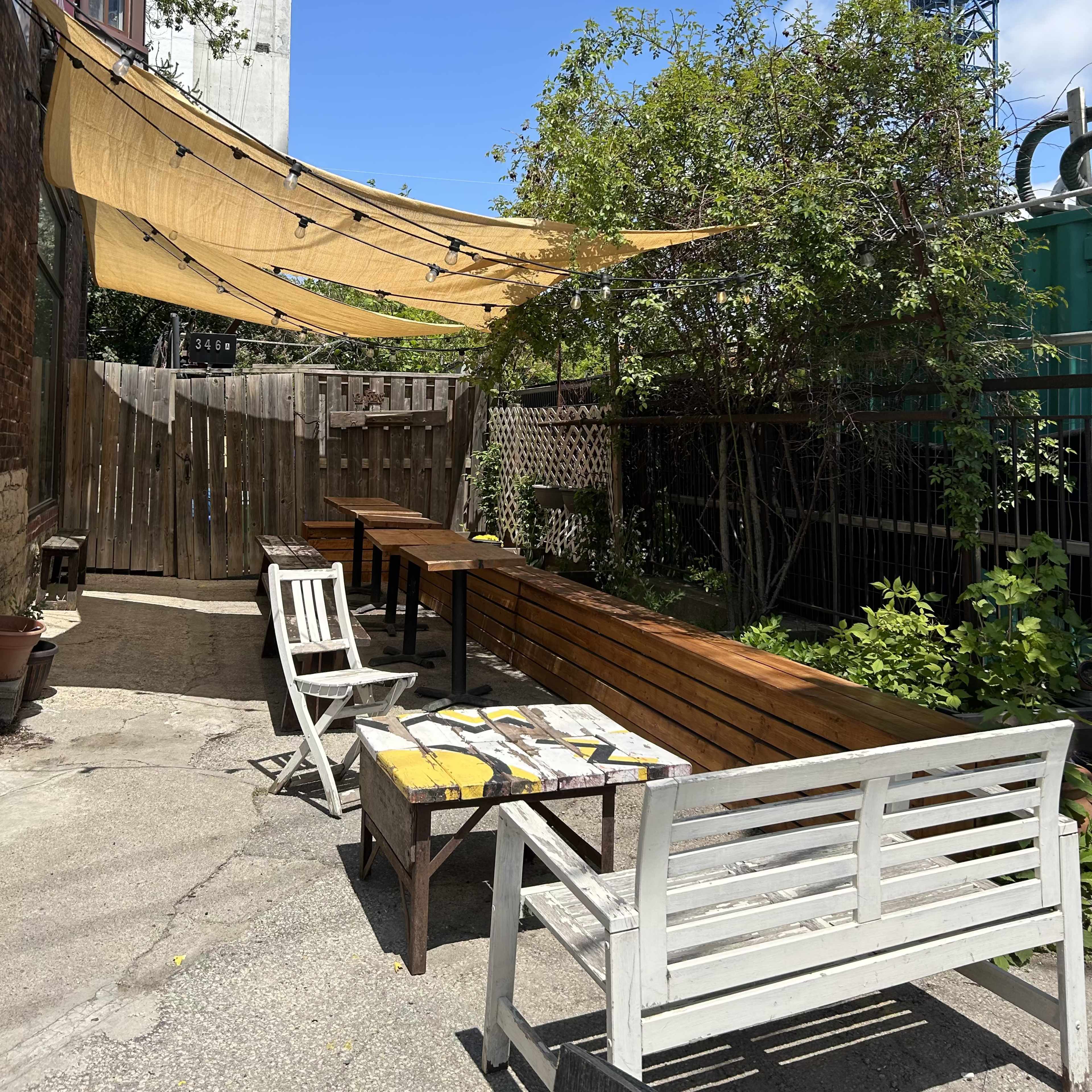 A patio area with wooden benches, tables, and a canopy overhead, surrounded by greenery and a construction site in the background.